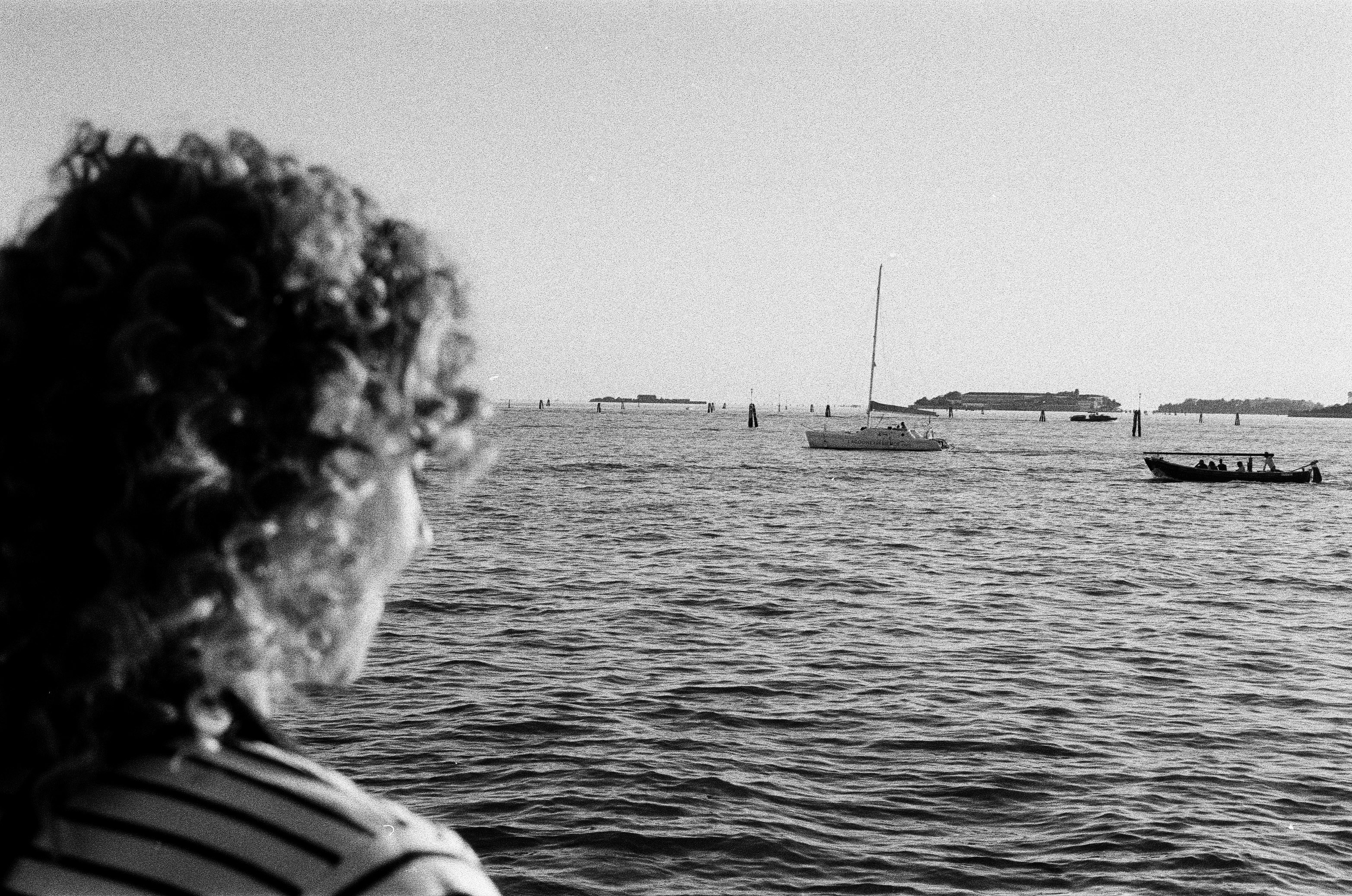 A person with curly hair looking out at a sailboat and boat on water