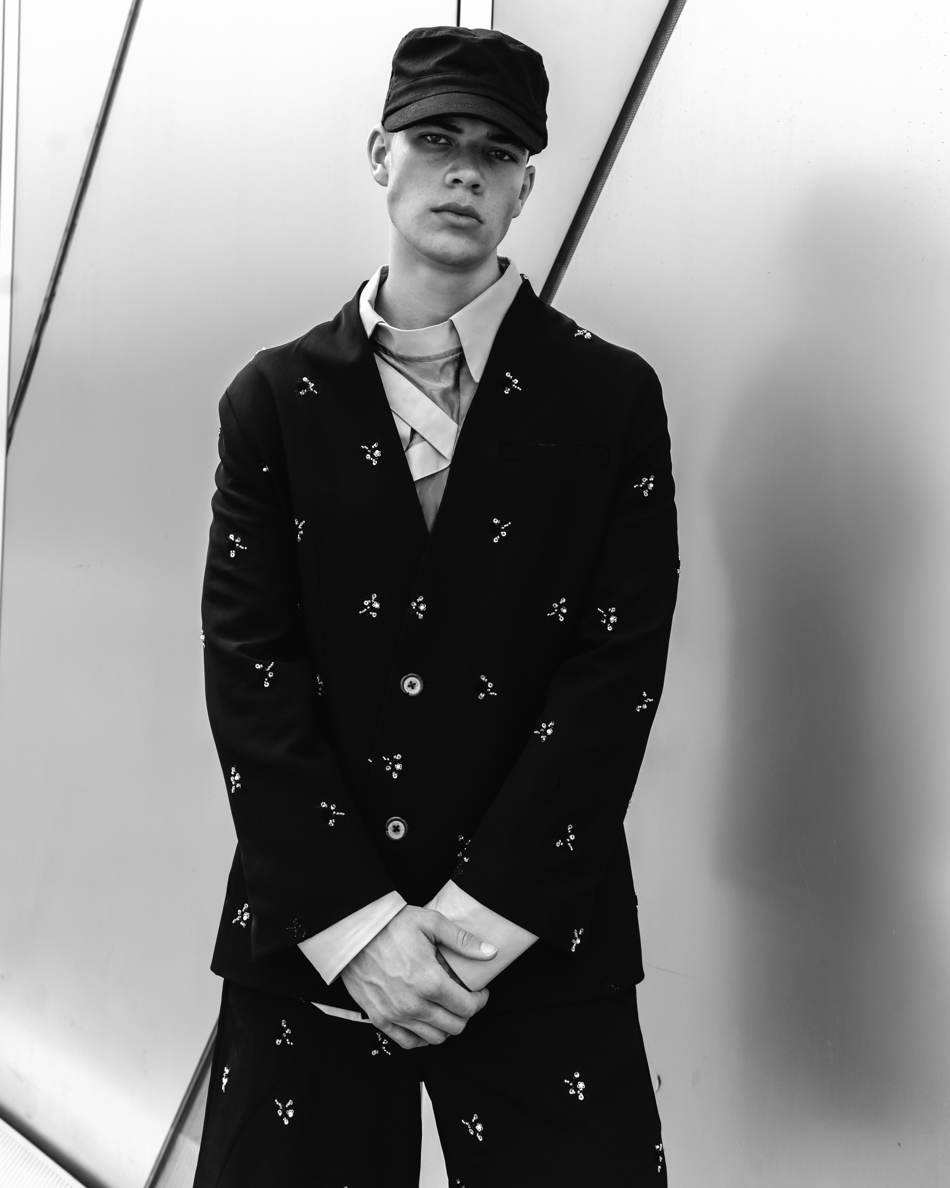 A person in a black embellished suit and cap against a smooth wall