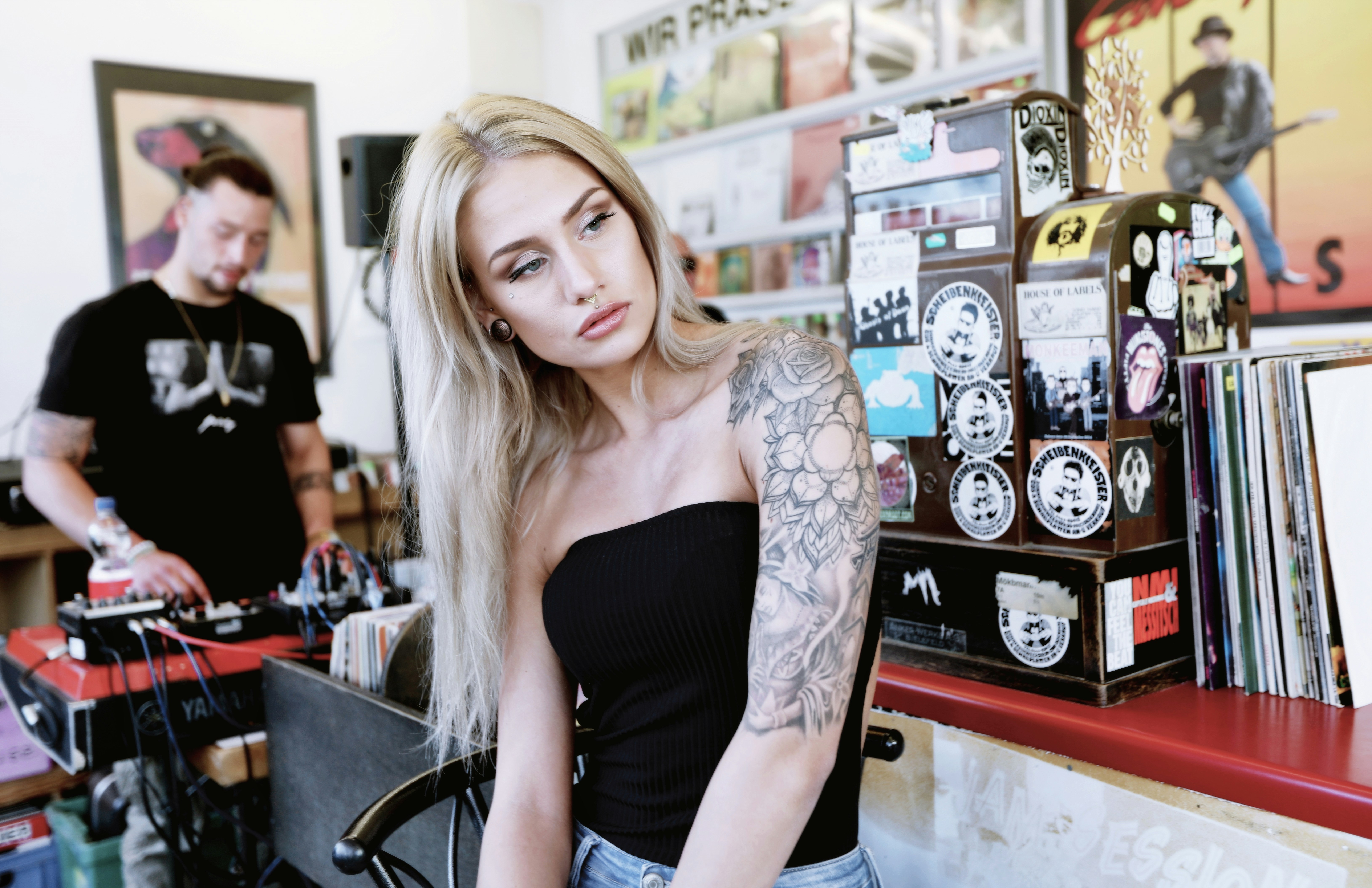 A blonde woman with a tattooed arm sitting near a sticker-covered cash register