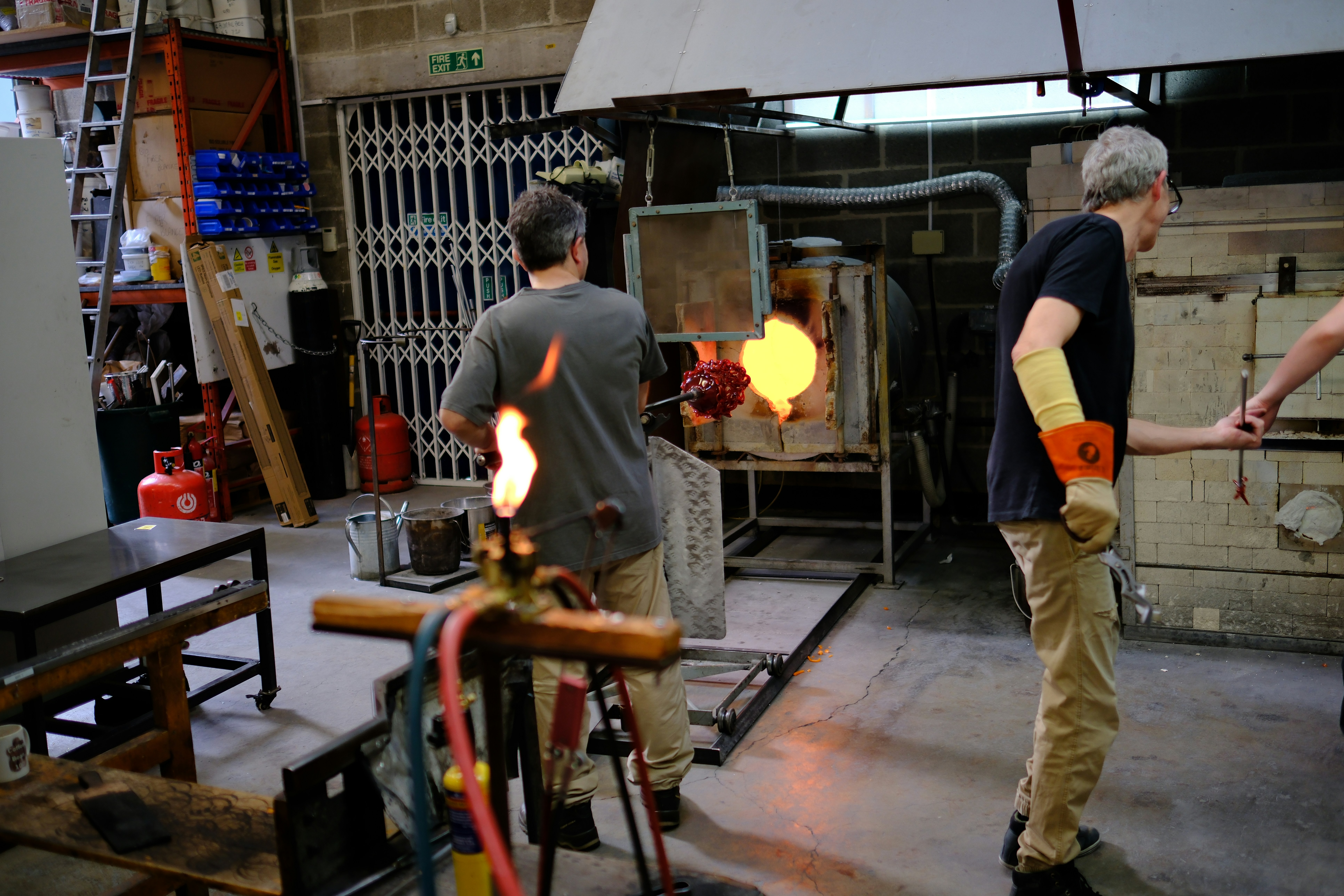 Two people working with molten glass in a workshop with a glowing furnace