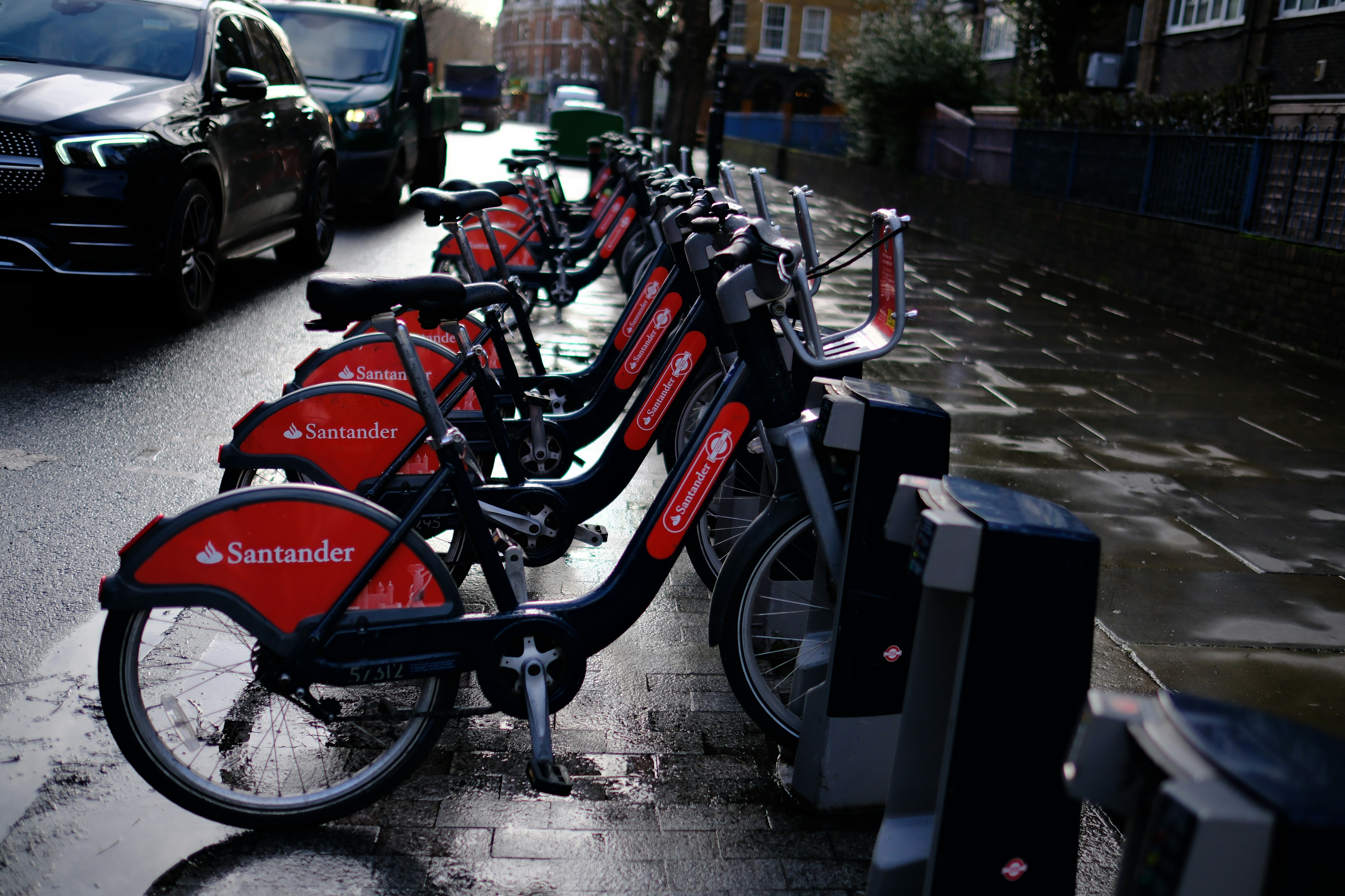 A row of red and black rental bicycles parked on a wet city sidewalk