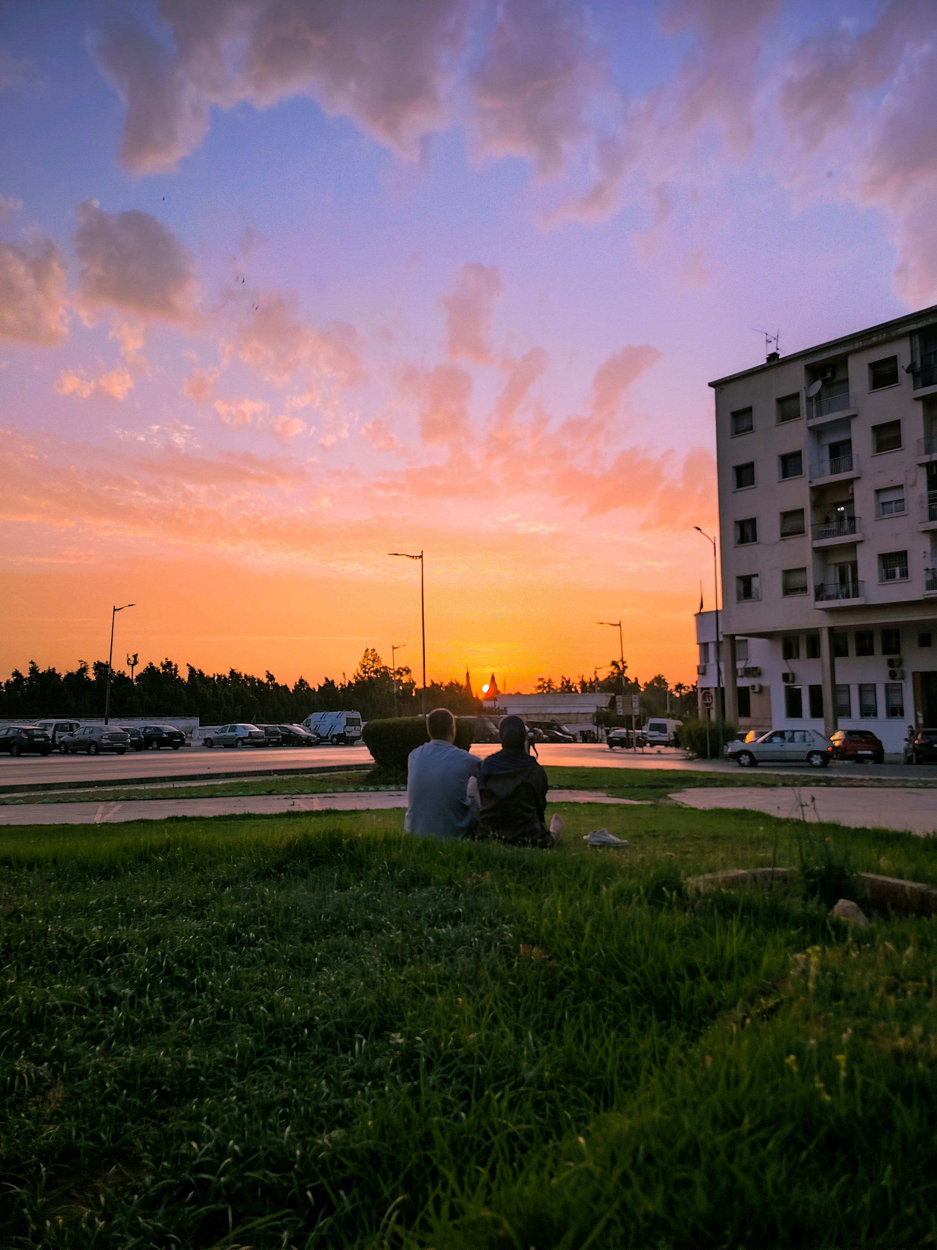 Two people sitting on grass in a parking lot under a warm orange sky