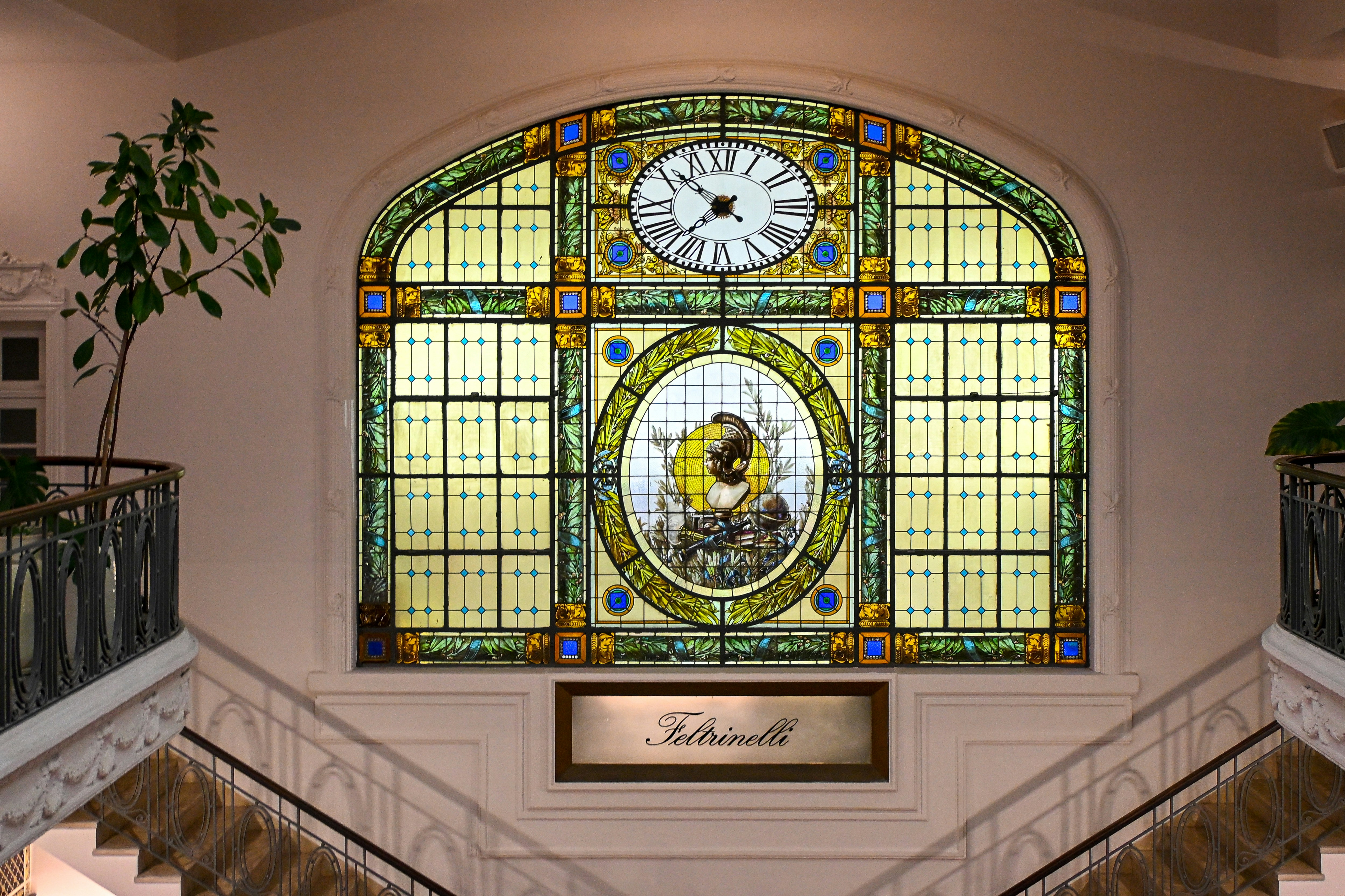 A large arched stained glass window with a central clock and classical bust