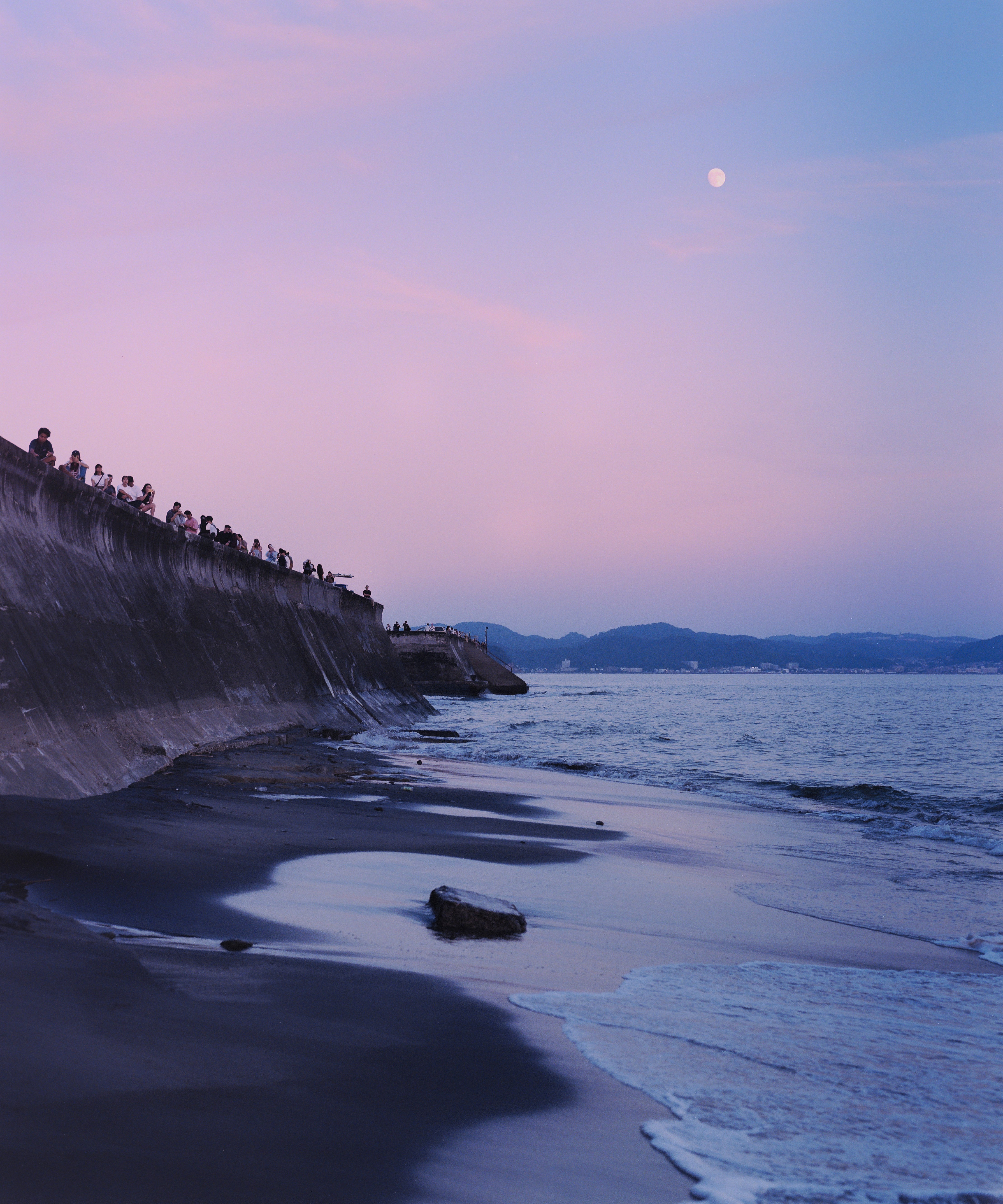 A group of people sitting on a concrete seawall above a dark sandy beach