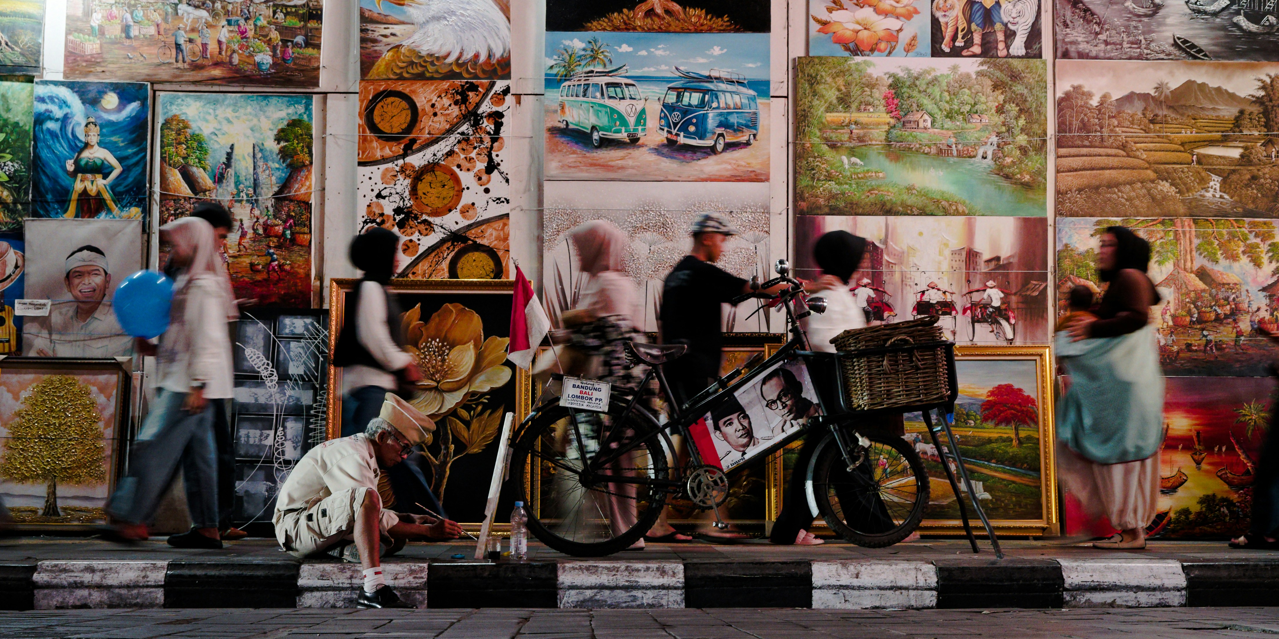 An artist painting on a sidewalk in front of a wall of framed paintings