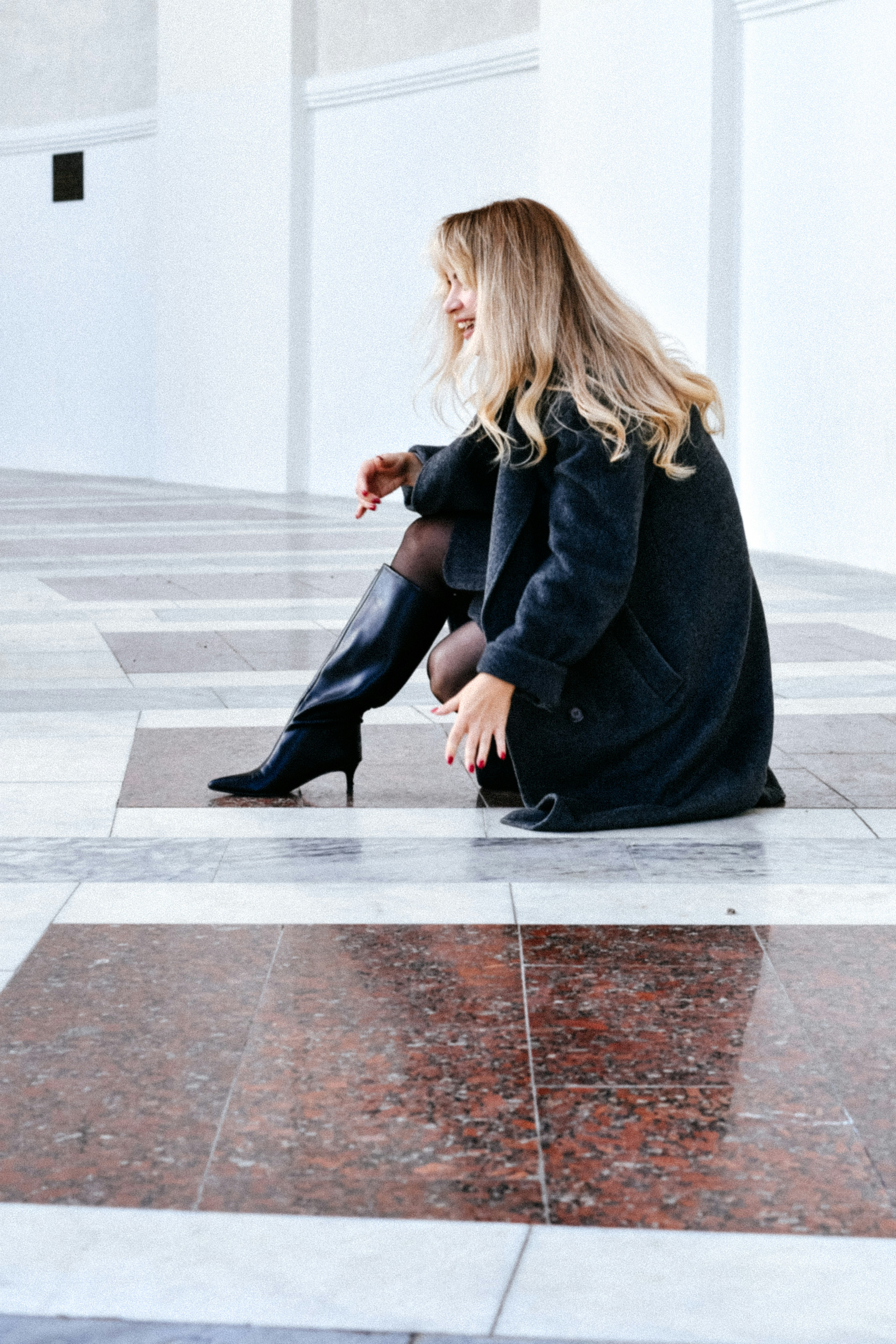 A woman with blonde hair in a dark coat and black boots crouching