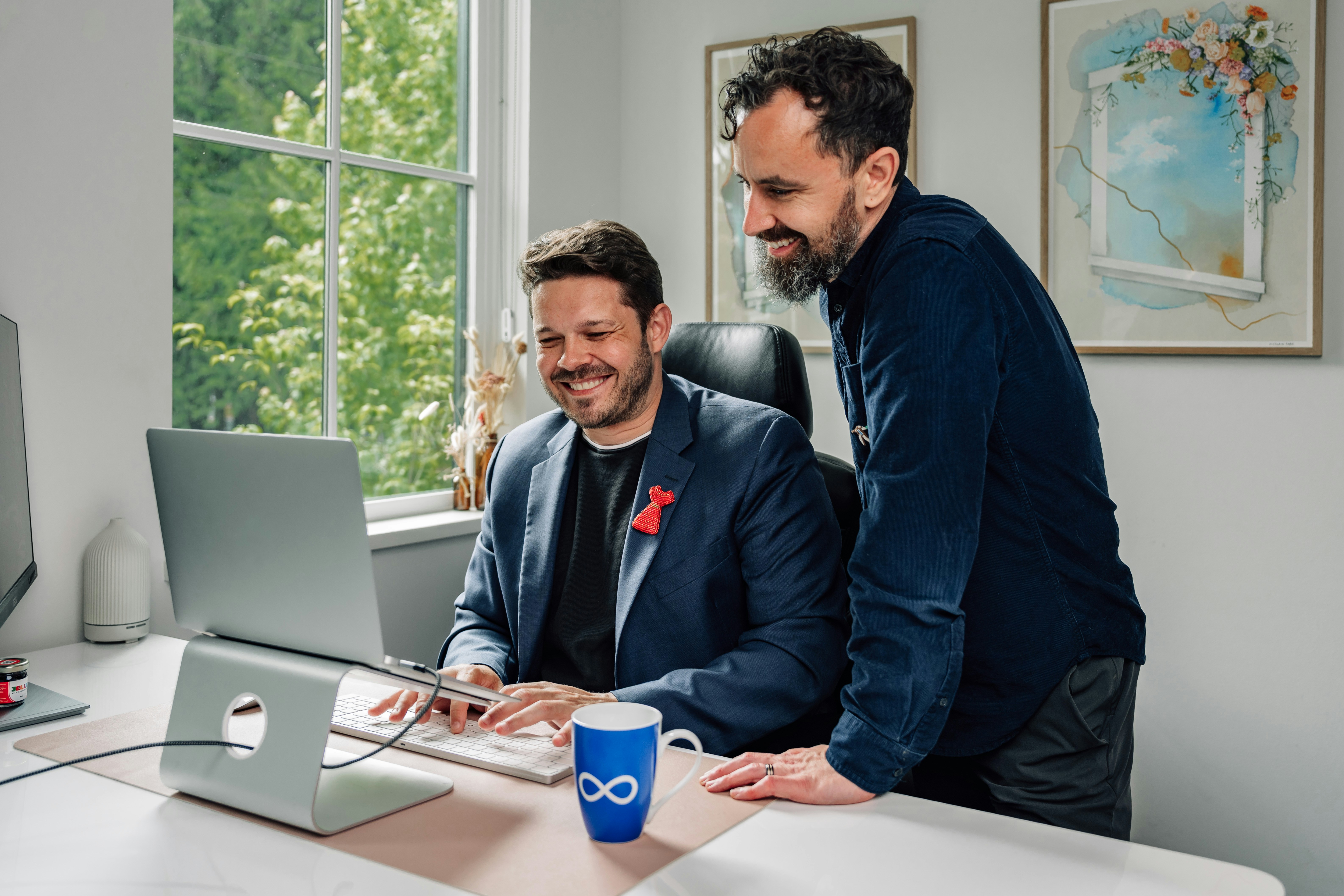 Two men smiling at a laptop on a desk in a bright office