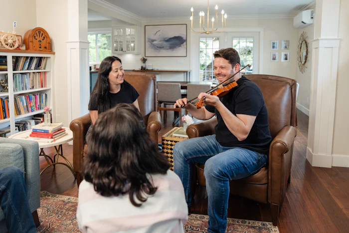 Un homme joue du violon dans un salon, devant une femme souriante et une fillette assise de dos