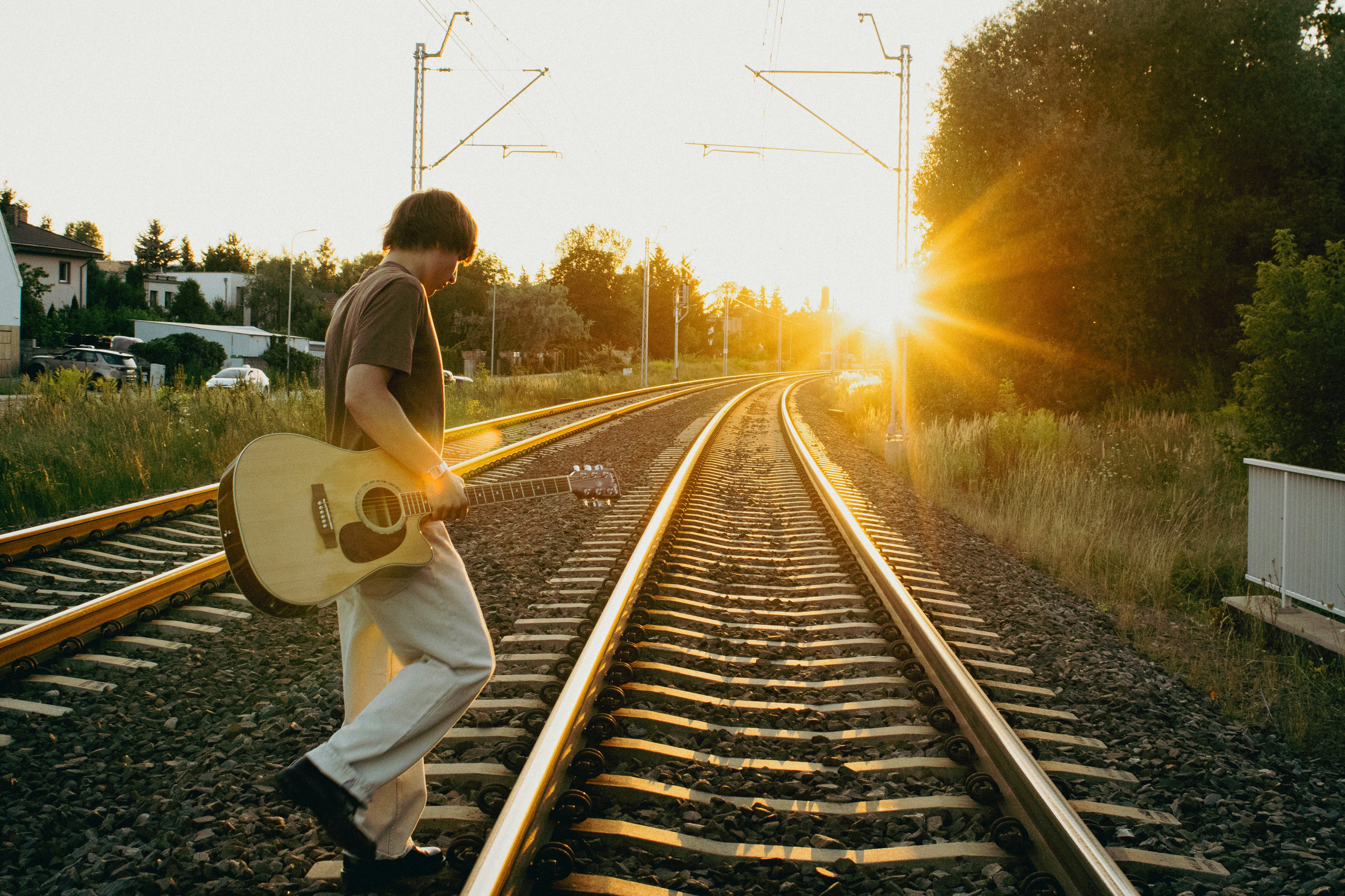 A person carrying an acoustic guitar walking along railway tracks in low golden light