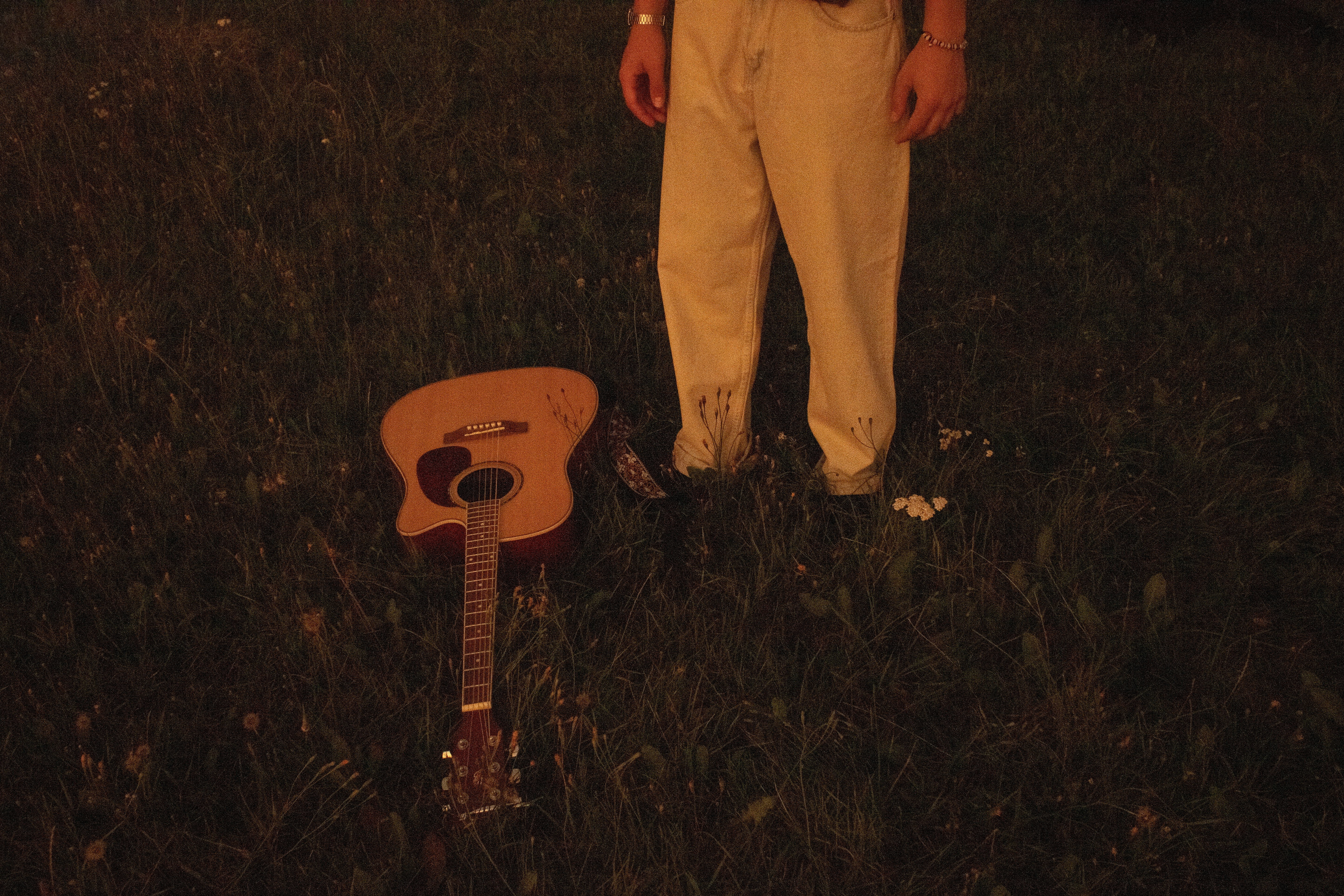 A person in light-colored trousers standing near an acoustic guitar in tall grass