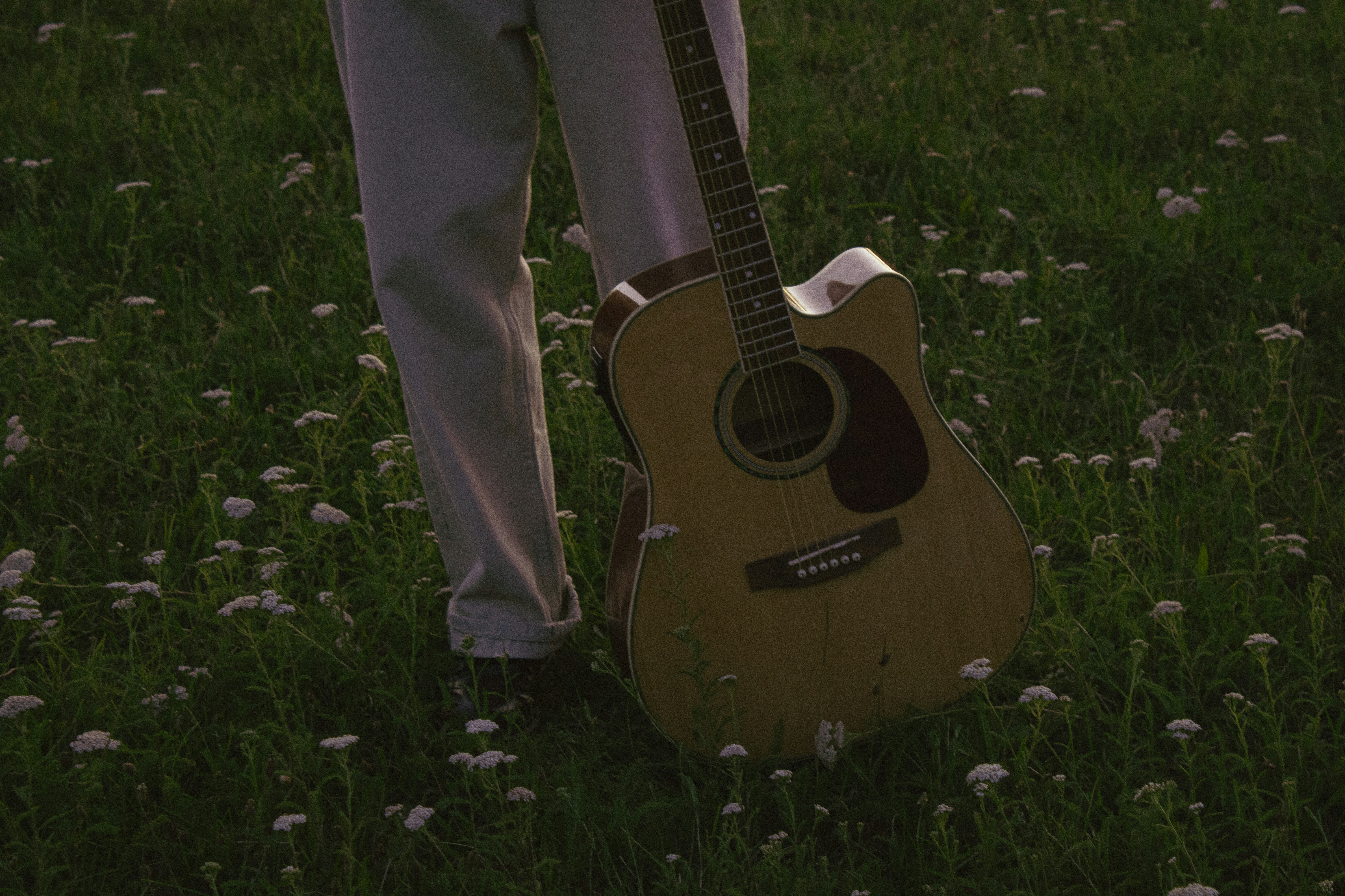 A person in light trousers standing with an acoustic guitar in a grassy field