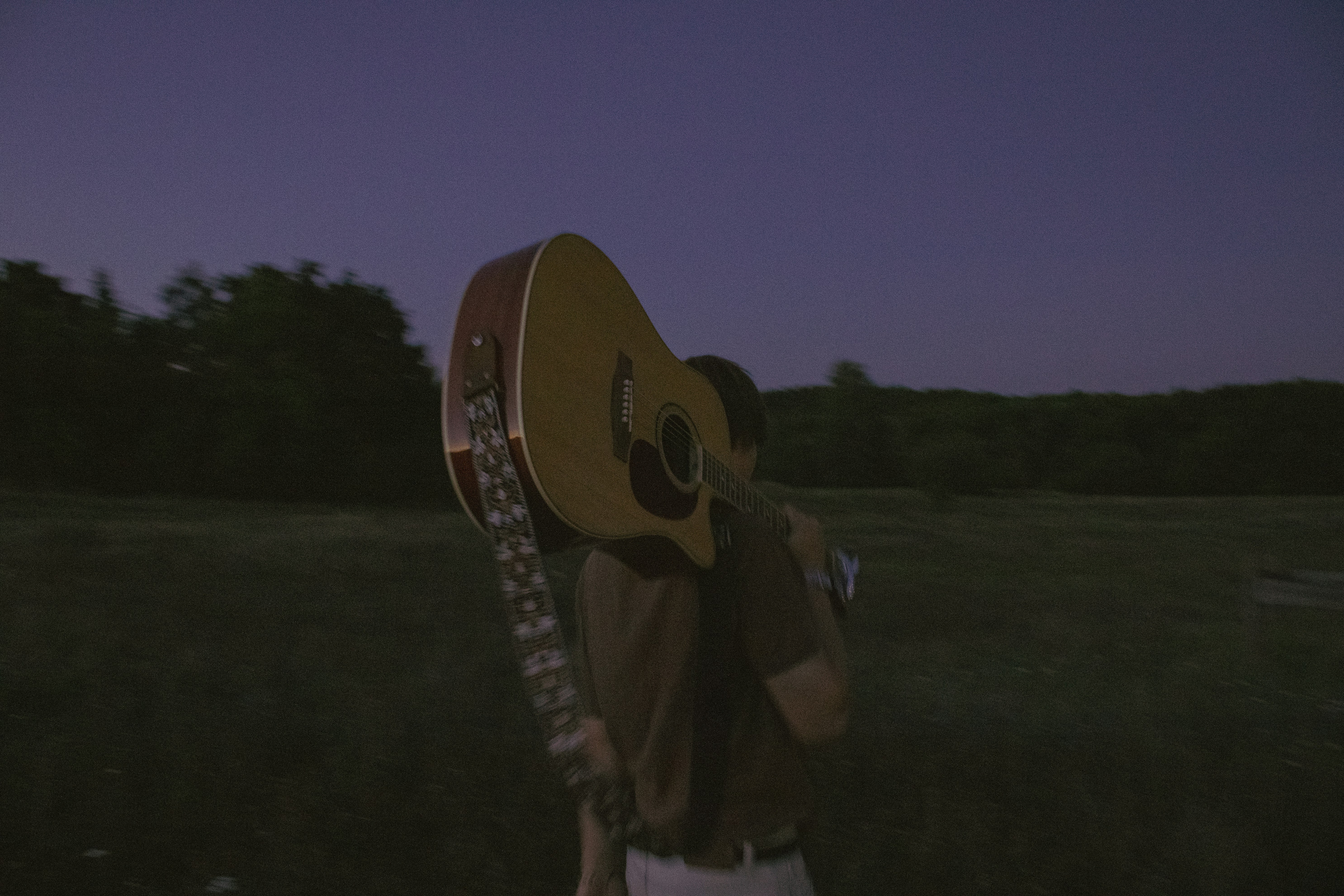 A person carrying an acoustic guitar on their shoulder across a dark field
