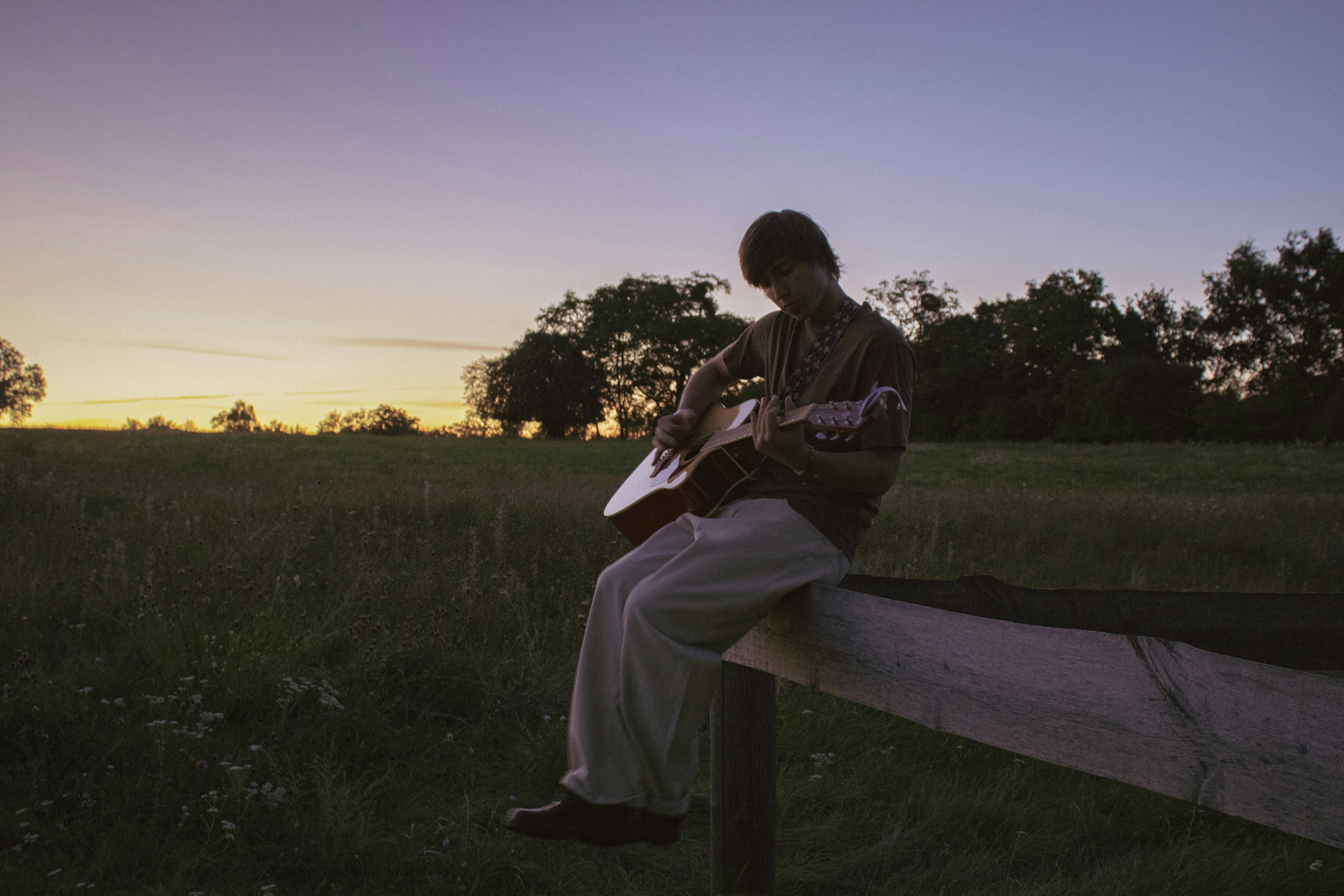 A person playing an acoustic guitar while sitting on a wooden fence at dusk