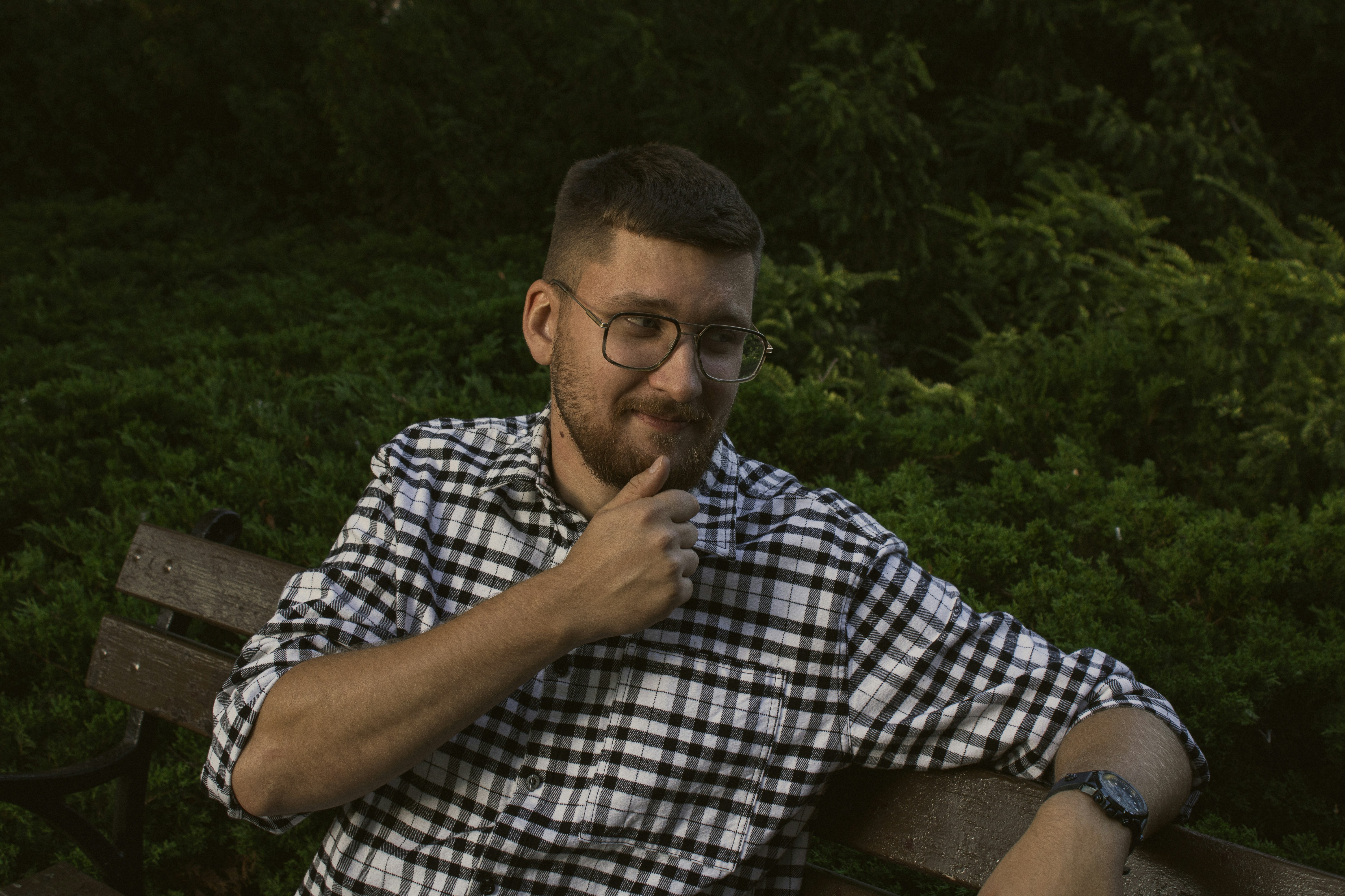 A man with a beard and glasses in a plaid shirt on a park bench
