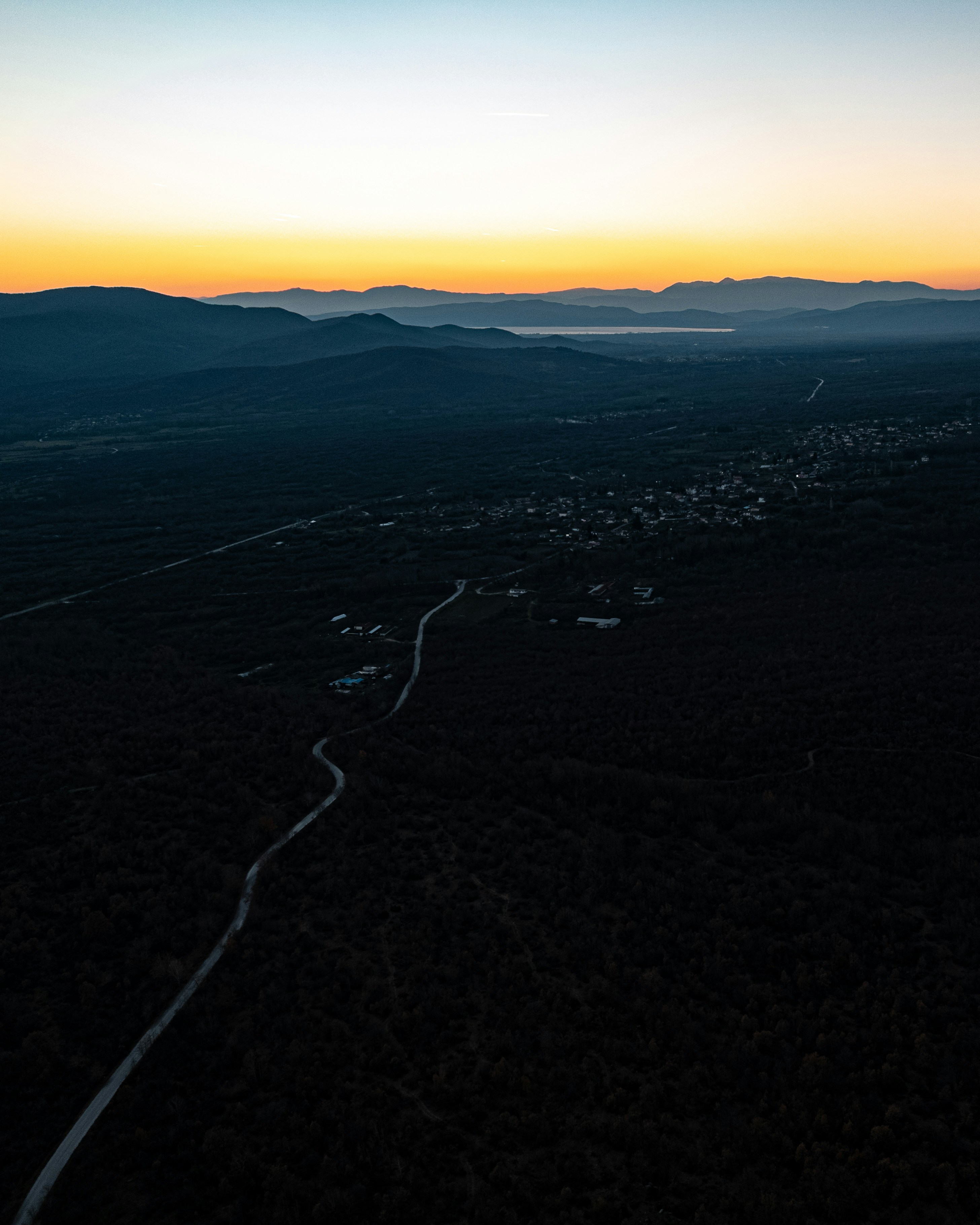 Aerial view of a winding road through a dark forest toward distant mountains
