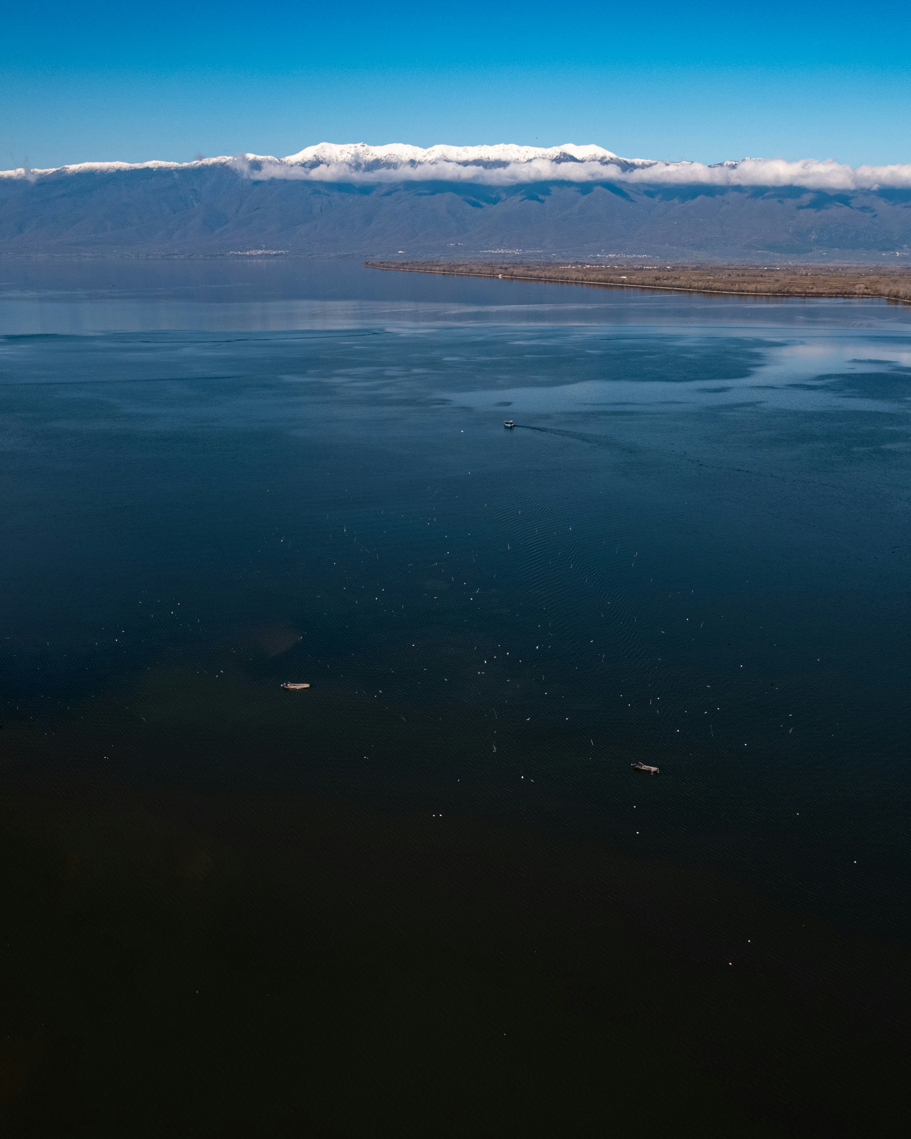 Small boats on a vast dark lake beneath snow-capped mountains under a clear sky