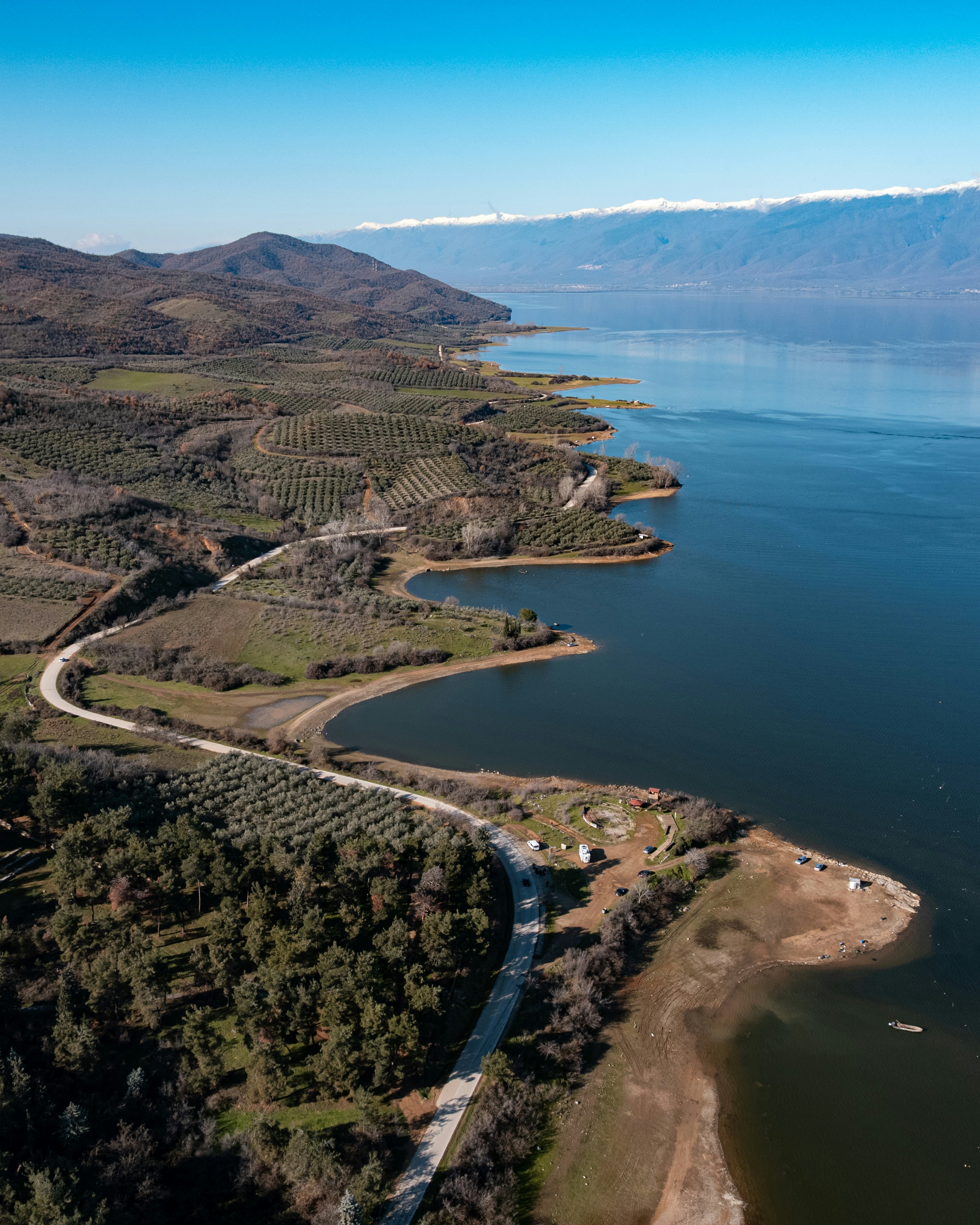 Aerial view of a winding road along a lake with snow-capped mountains