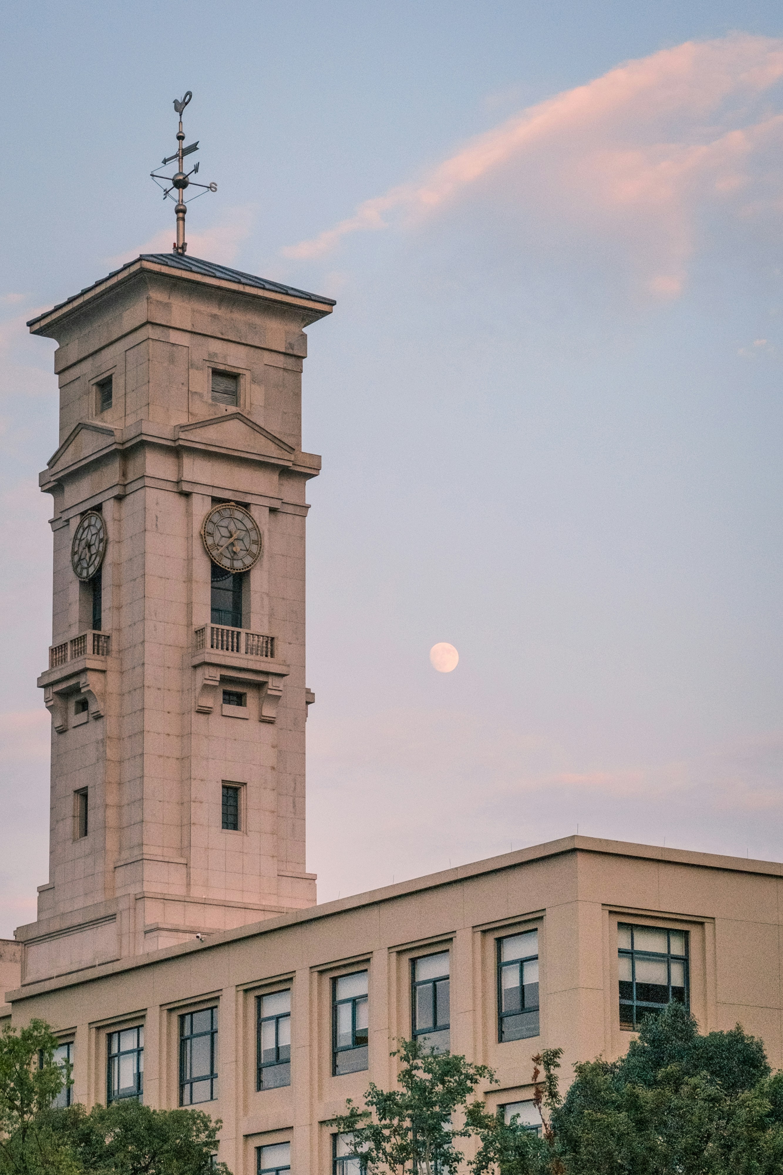 A stone clock tower with a weather vane against a pale sky with the moon