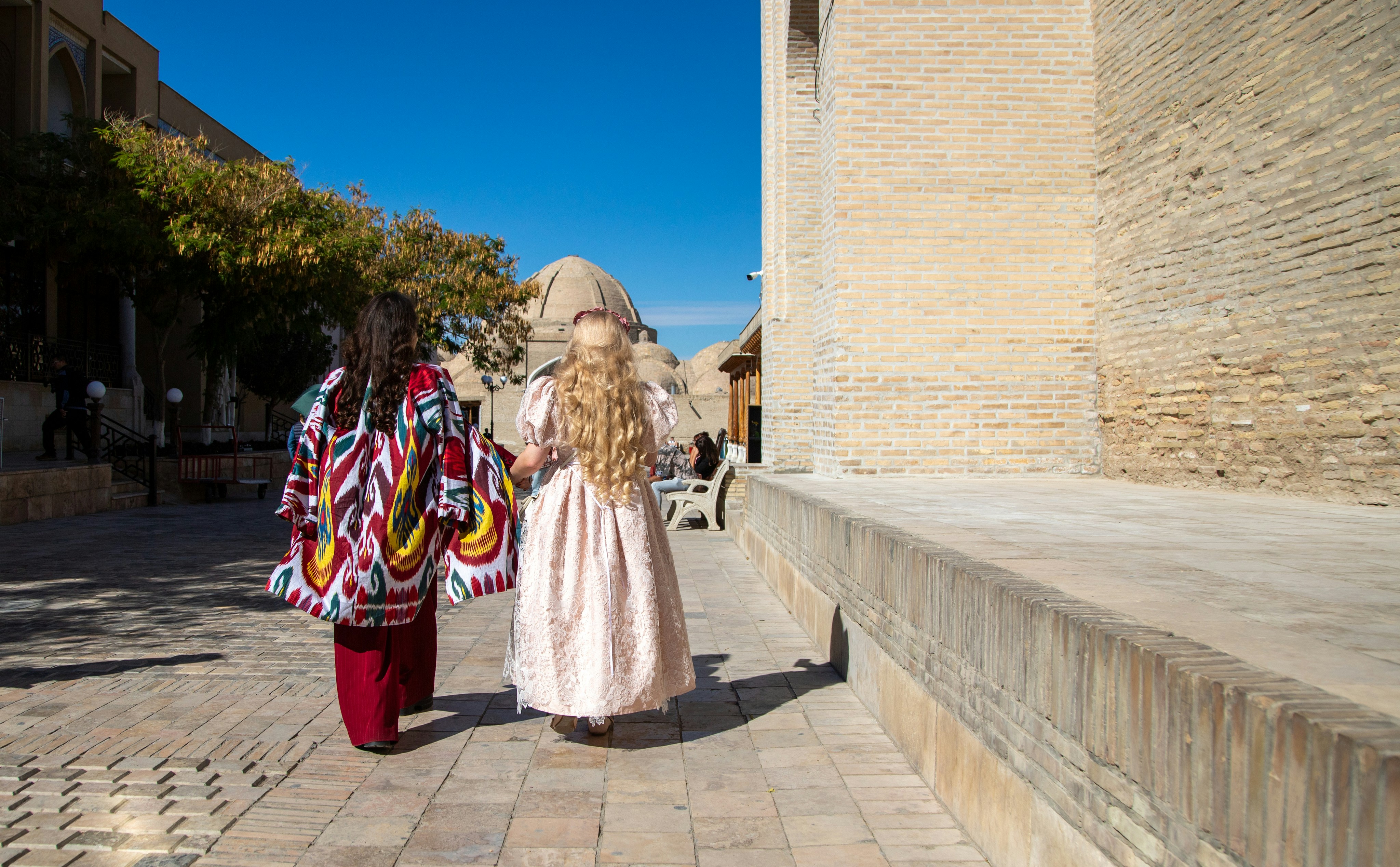 Two women walking on a stone path beside a brick wall in Bukhara