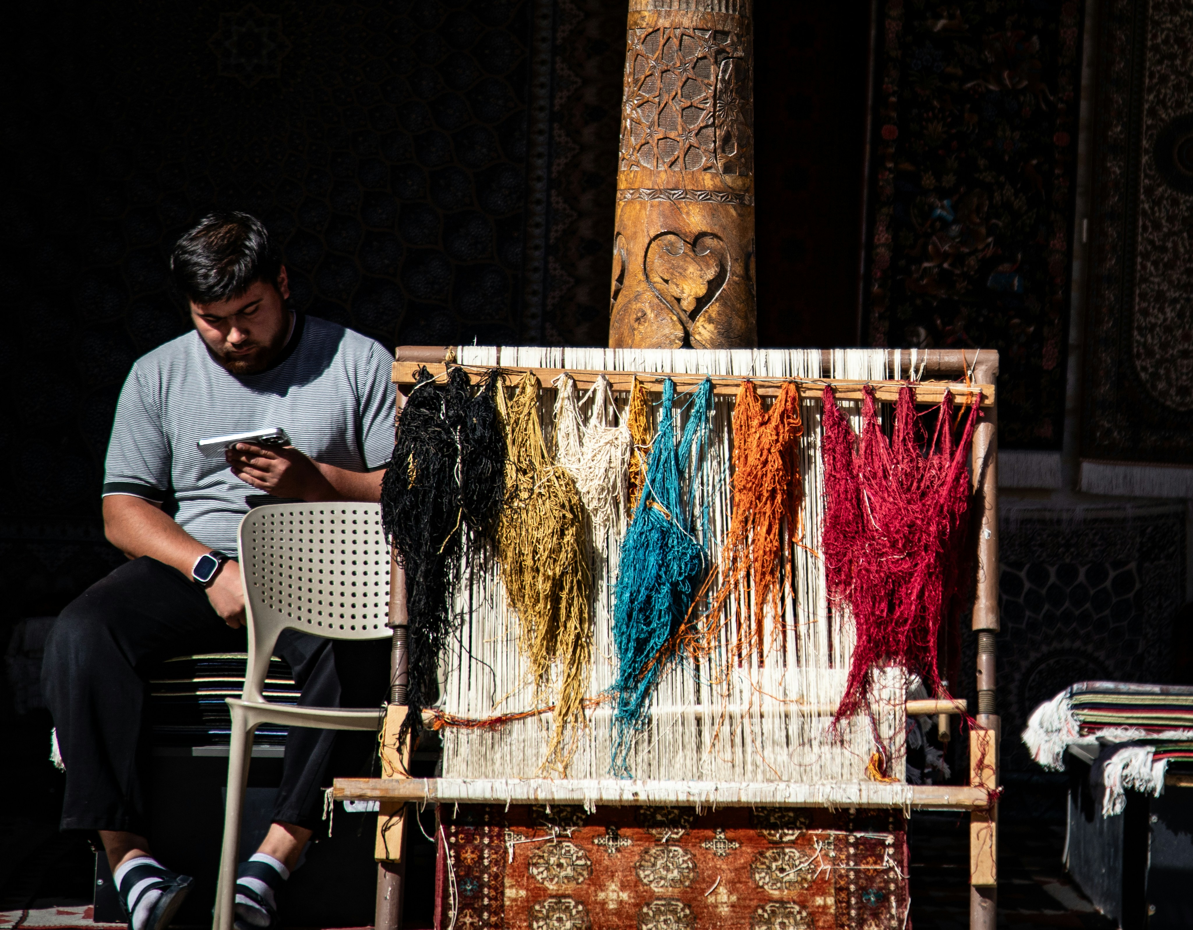 A person using a smartphone beside a wooden loom with colorful hanging yarn