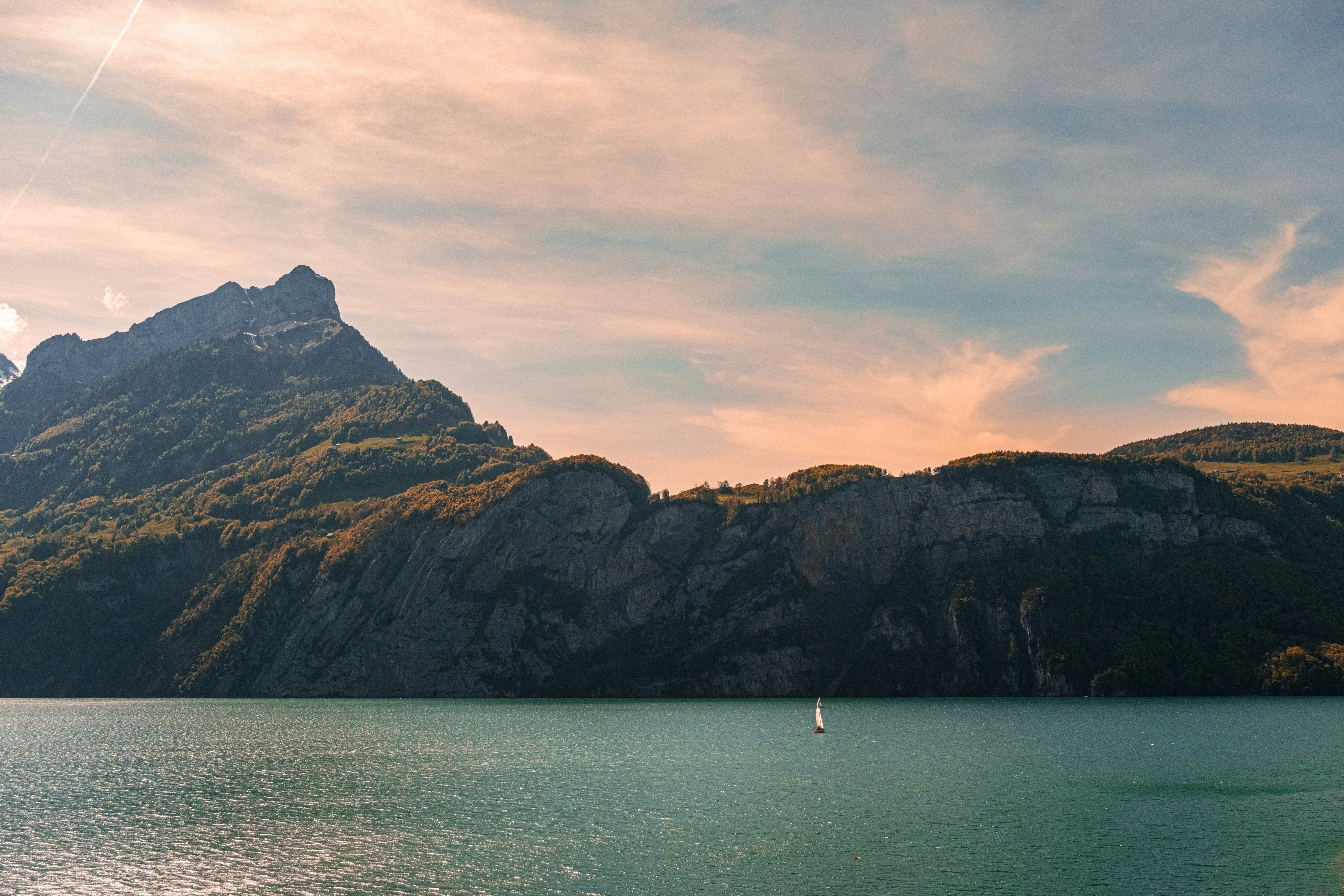 A small sailboat on a vast lake beneath towering mountains under a warm sky