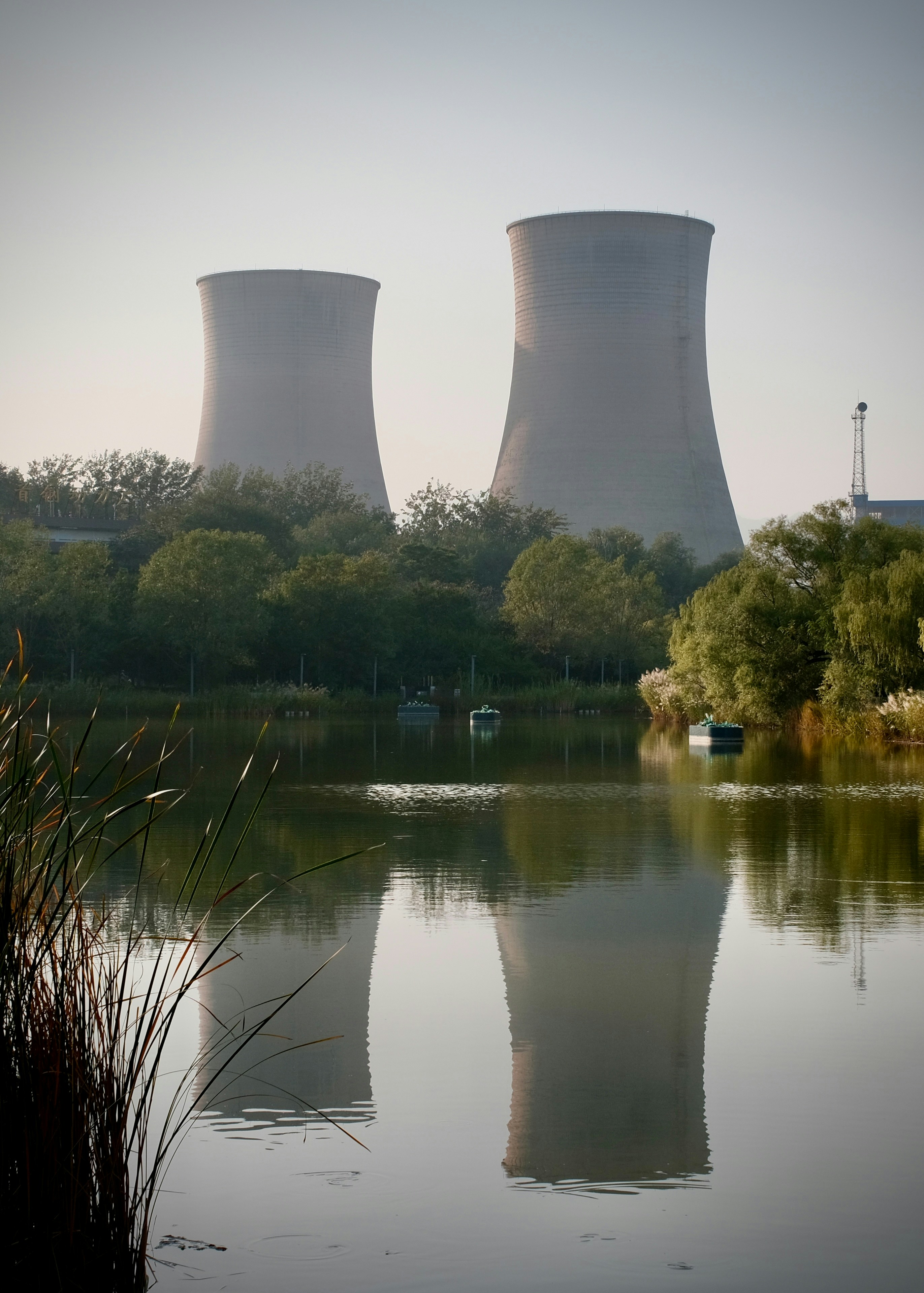 Two large cooling towers reflecting in a calm lake surrounded by green trees