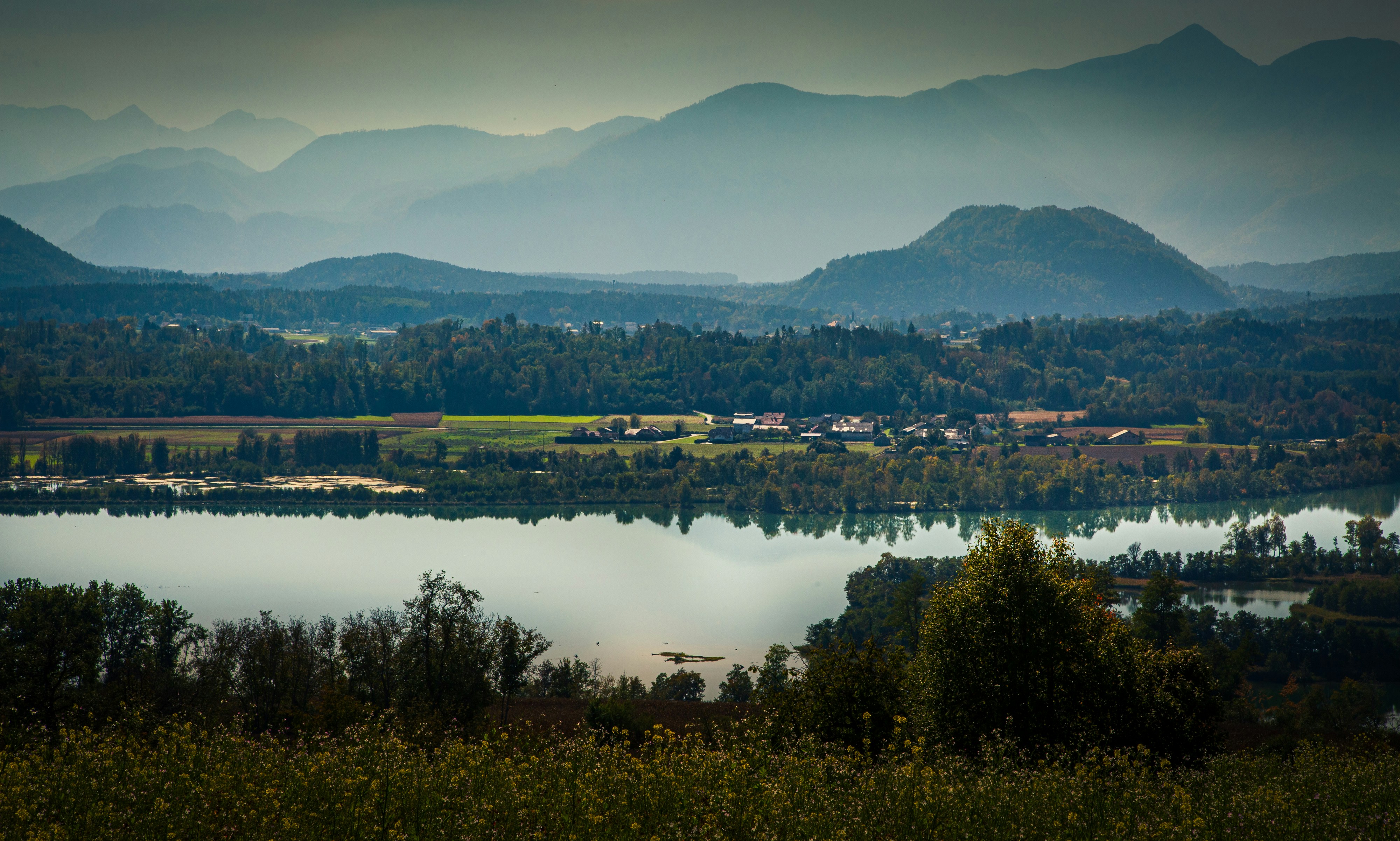 A calm lake reflecting distant mountains and a small village nestled in a green valley