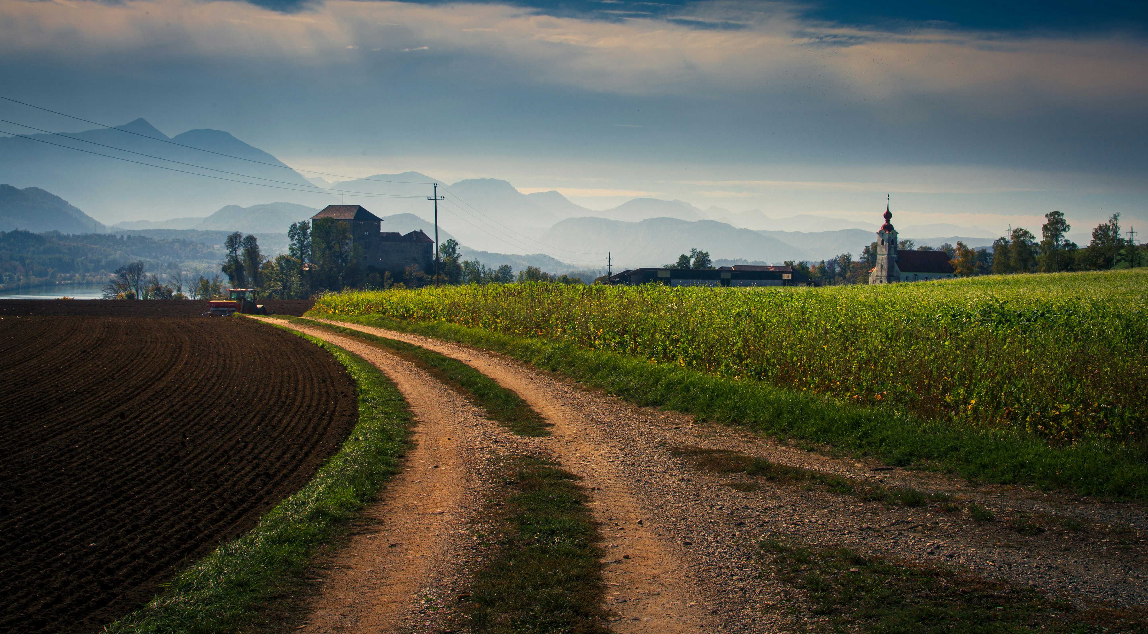 A dirt road winding through fields toward a castle and church under distant mountains