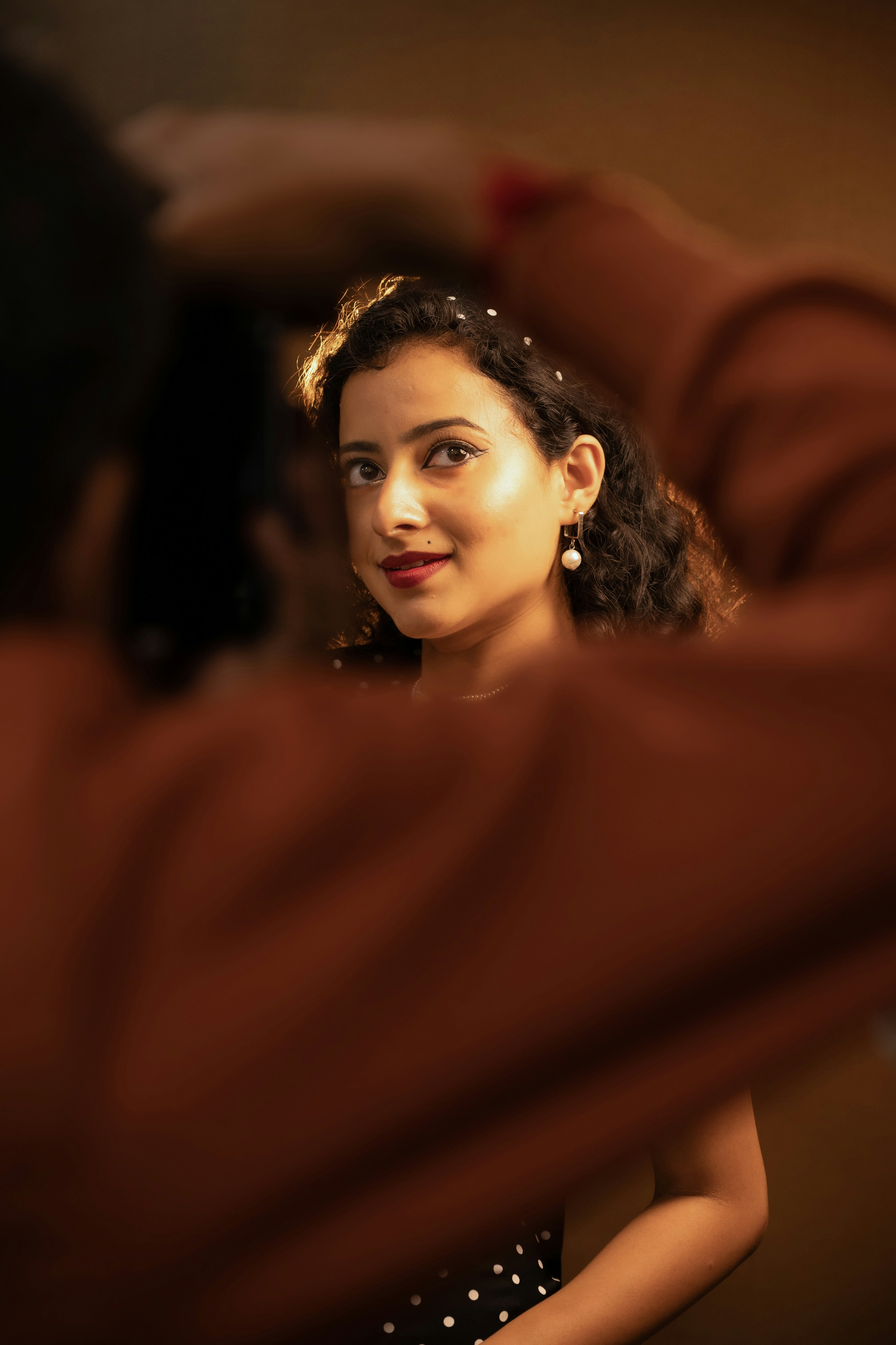 A woman with curly hair and pearl earrings looking toward a camera in warm light