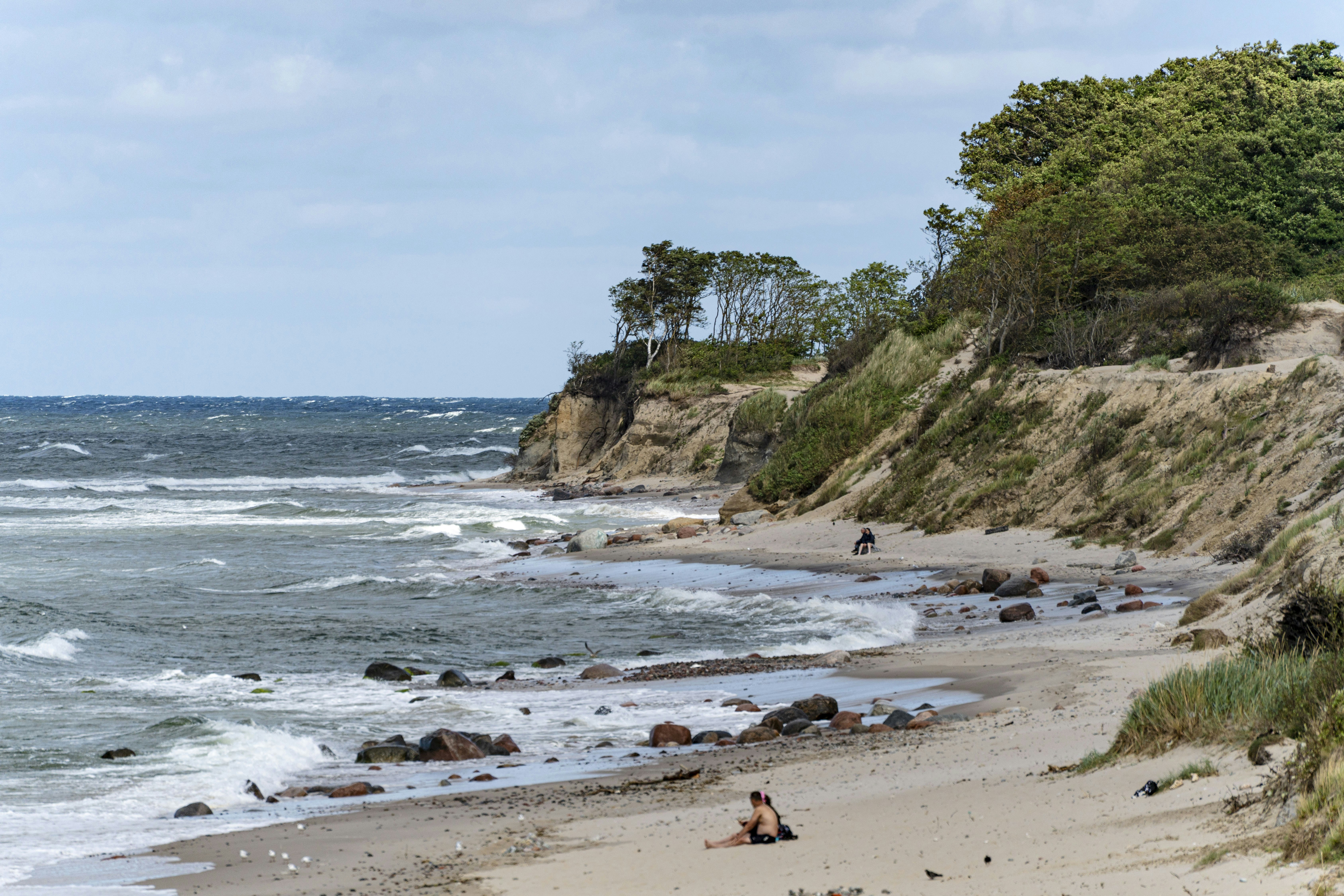 A sandy beach with rocky cliffs and trees under a cloudy sky