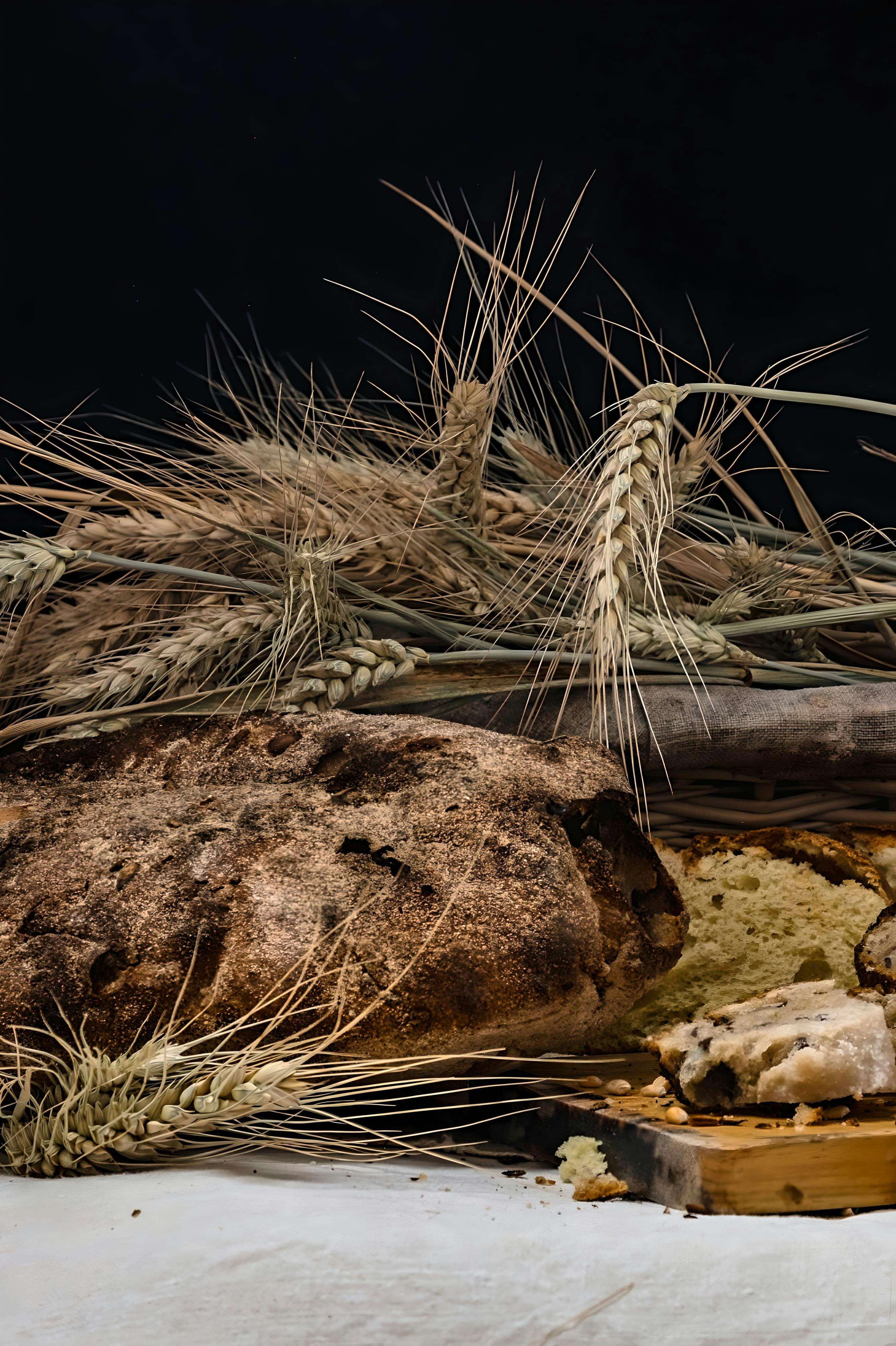 A rustic loaf of dark bread beside wheat stalks and a slice of bread on a wooden board