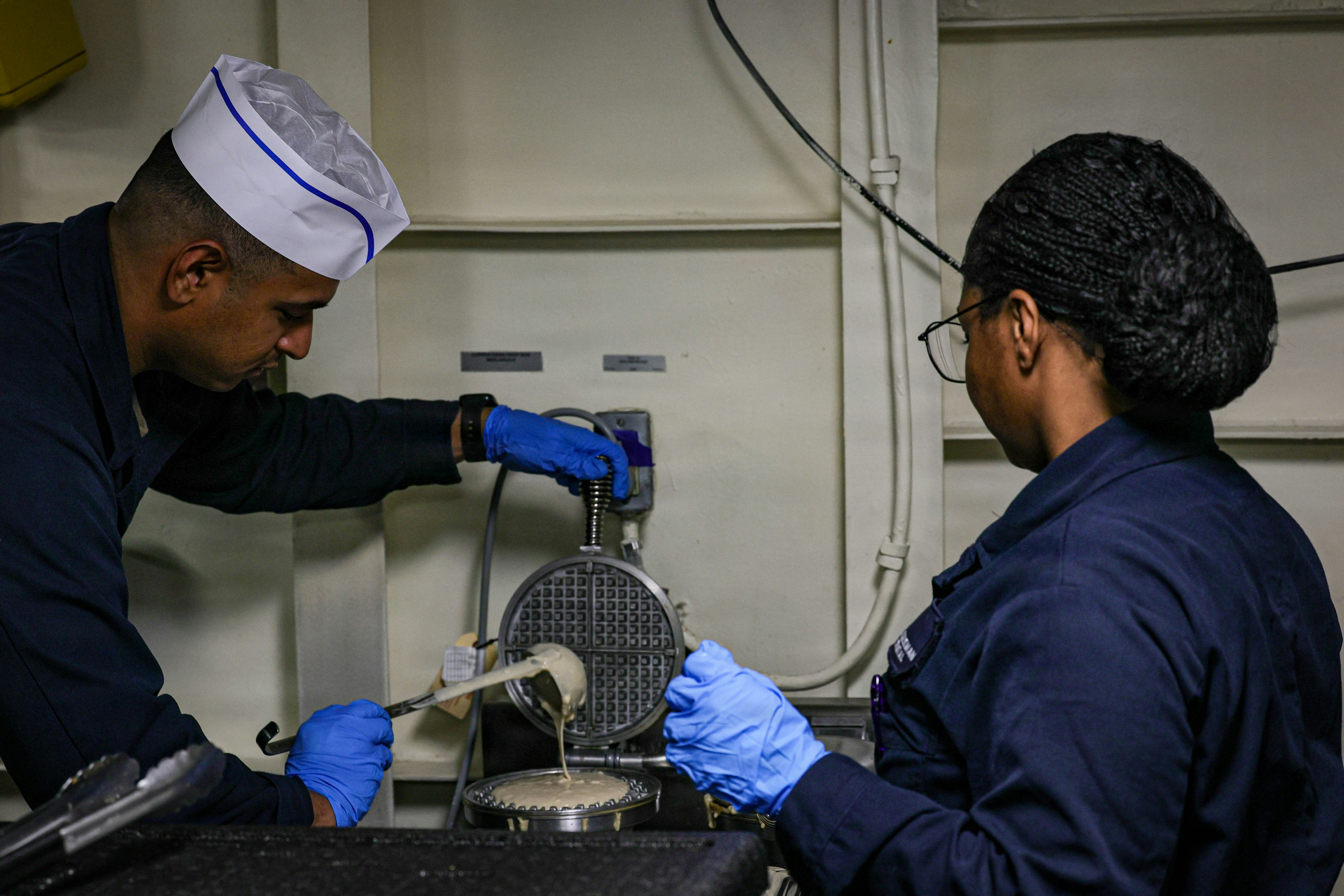 Two people in blue uniforms and gloves pouring batter into a waffle iron