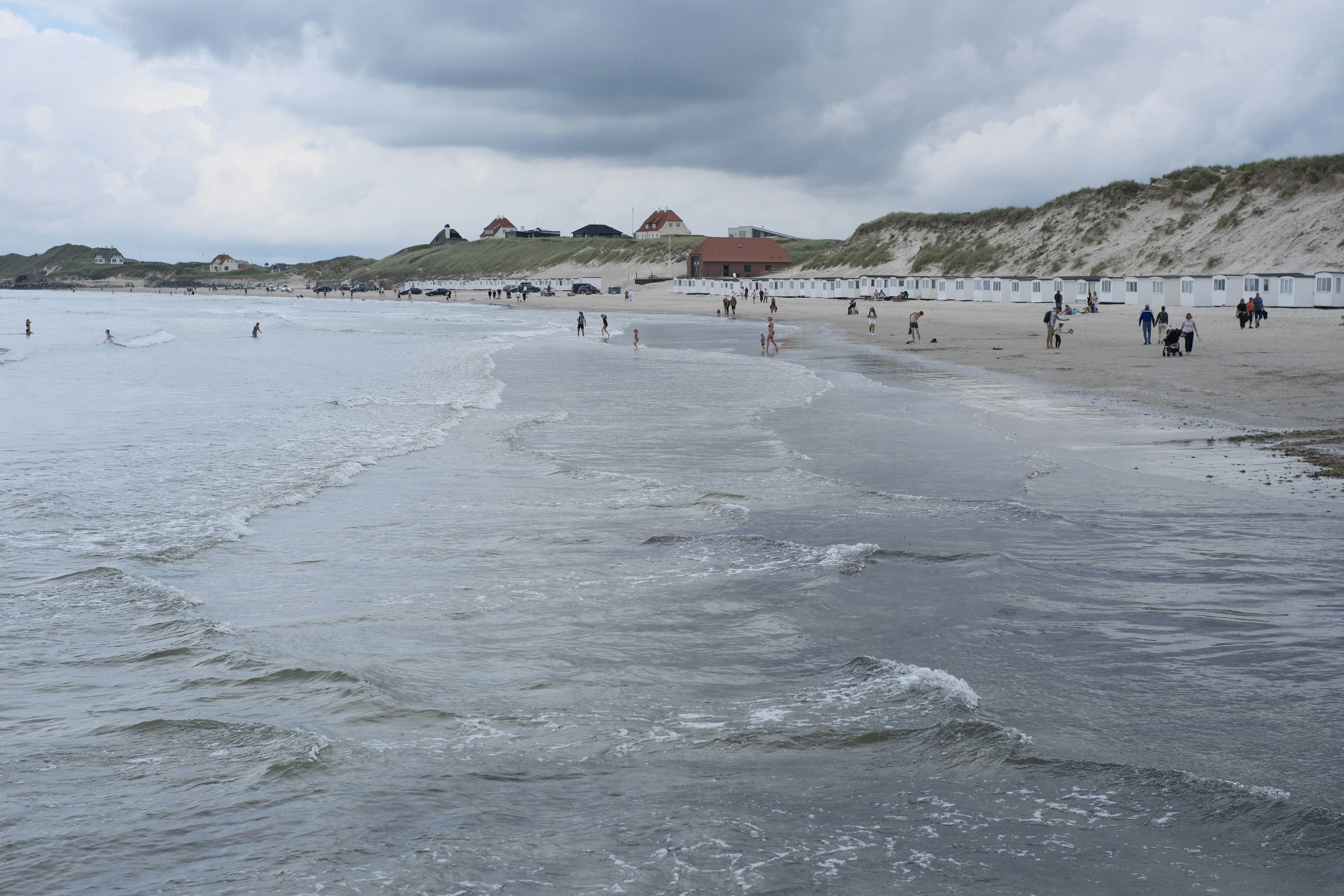 People on a sandy beach with small white cabins under a cloudy sky