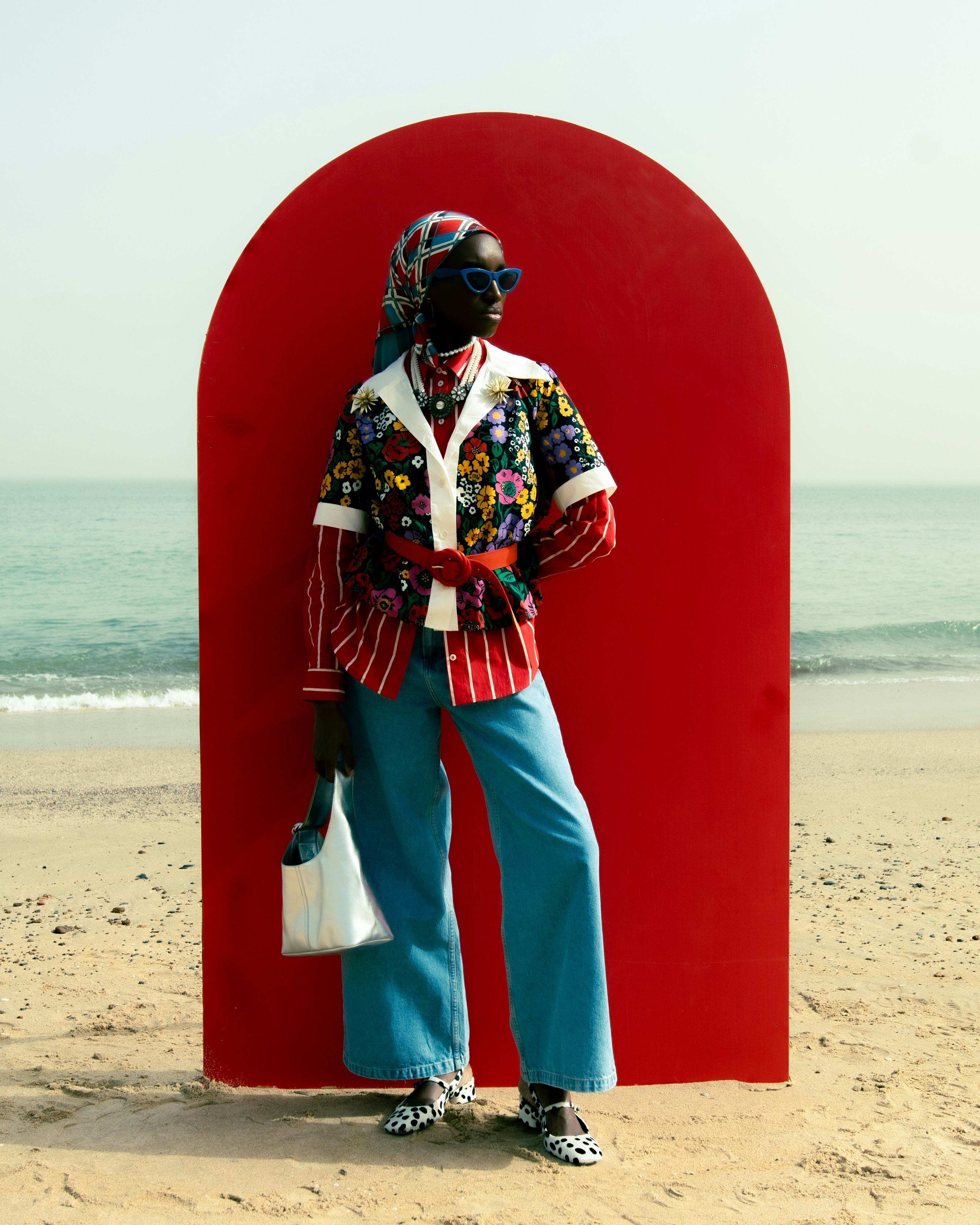 A person in a floral jacket and headscarf standing before a red arch on a beach