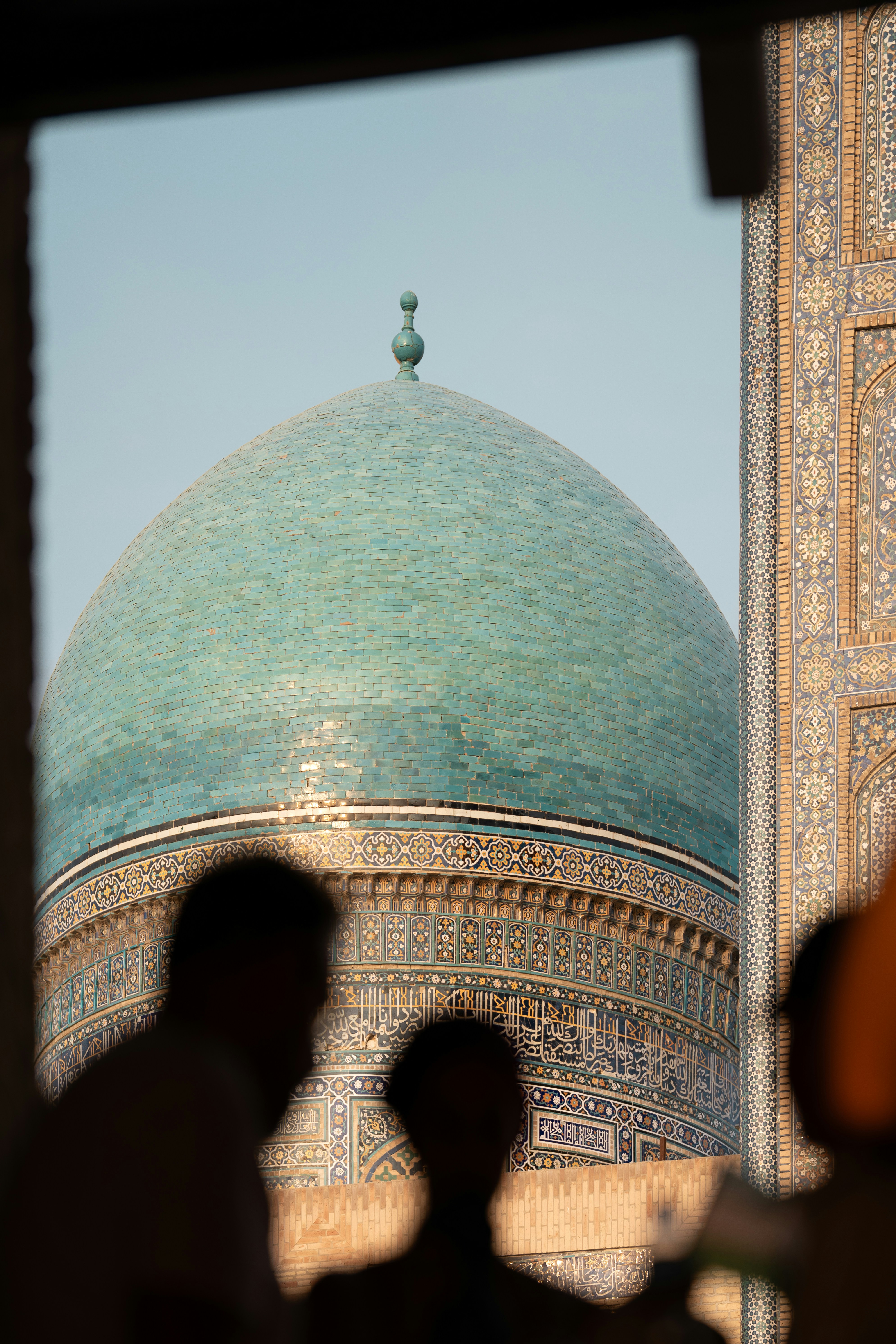 A turquoise tiled dome of a mosque in Bukhara, Uzbekistan, framed by silhouettes