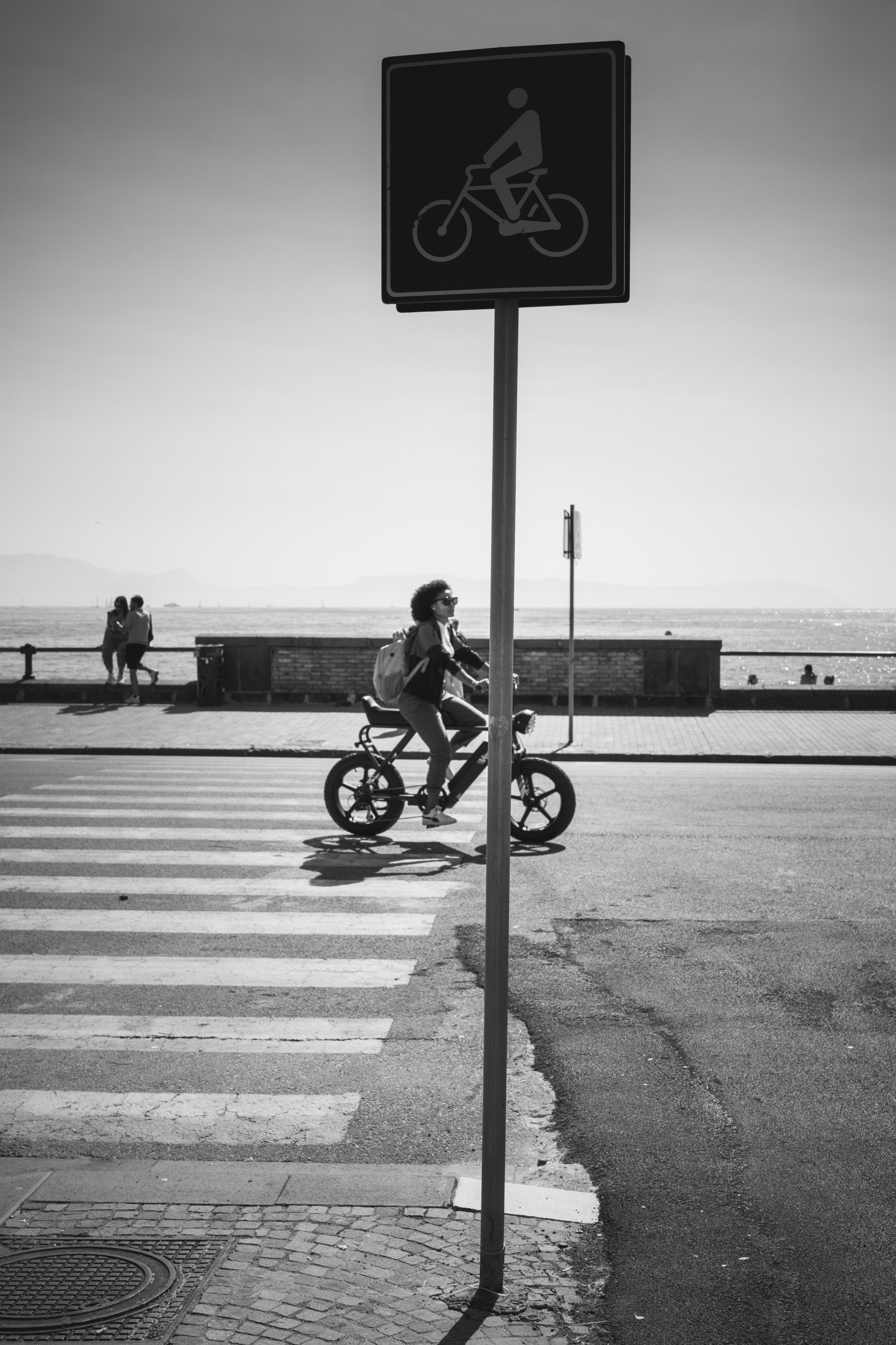 A person cycling an electric bike across a crosswalk near a coastal promenade