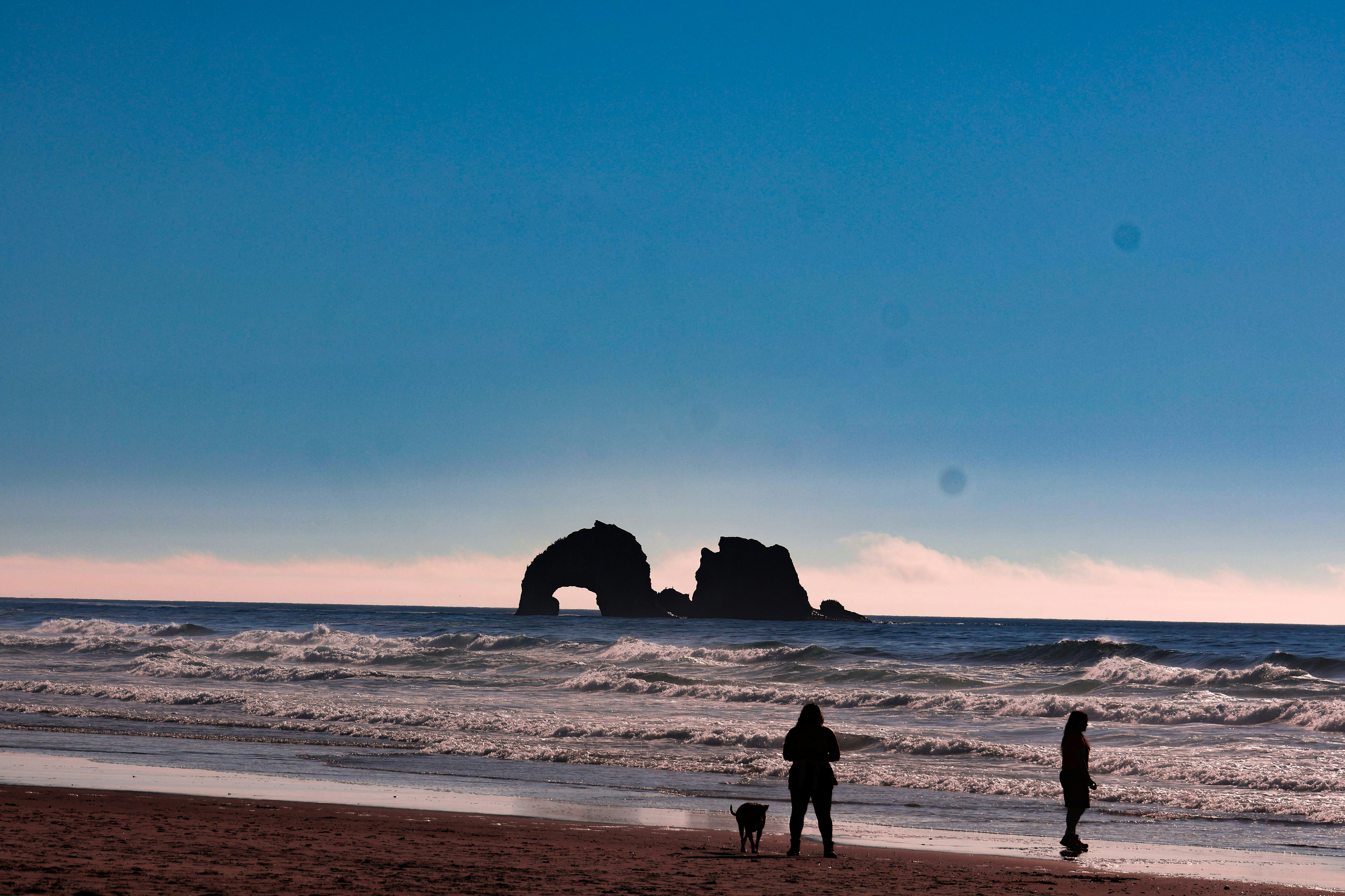 Two people in silhouette on a wet sand beach looking toward sea stacks under a blue sky