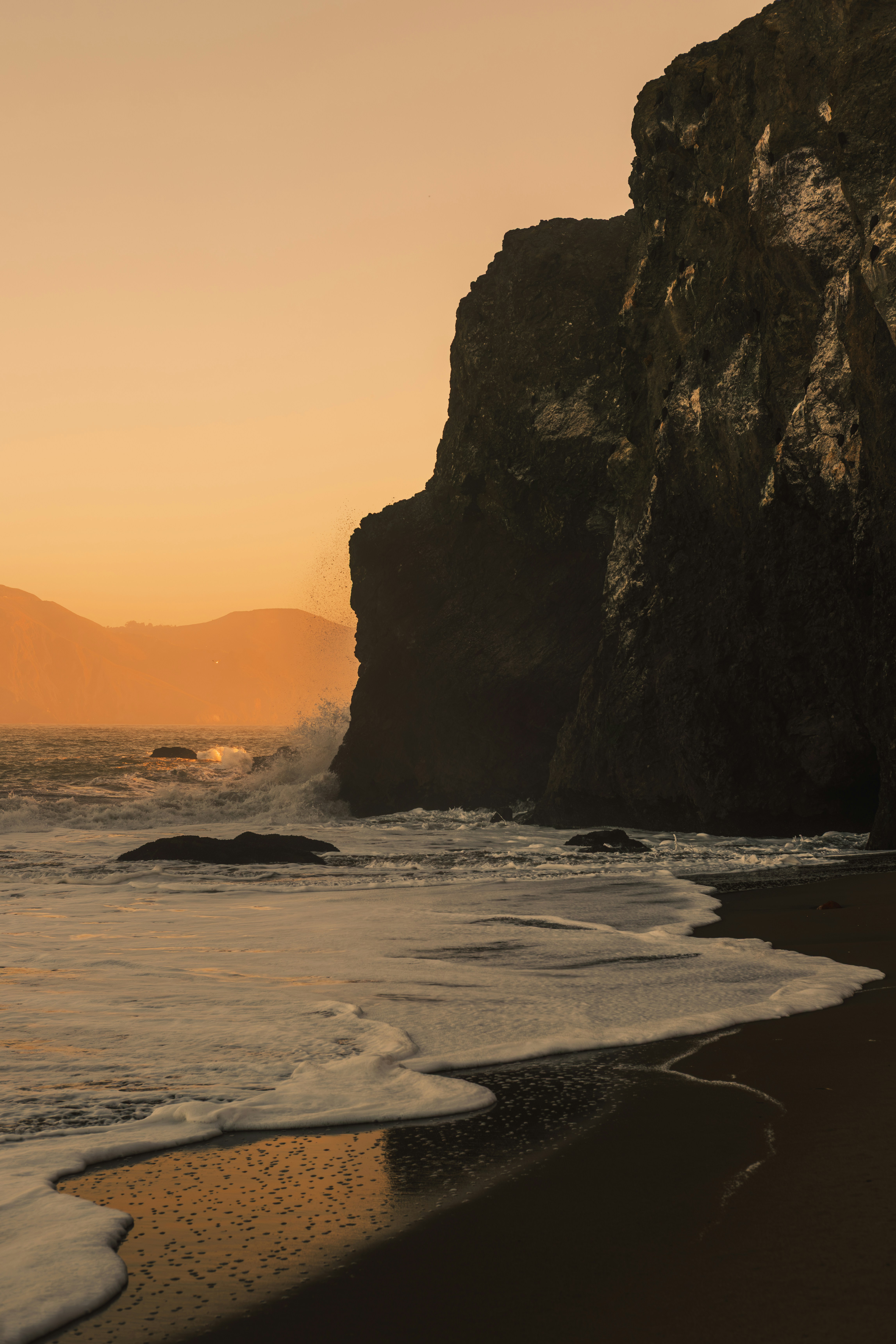 Waves crashing against a dark rocky cliff on a beach at golden hour