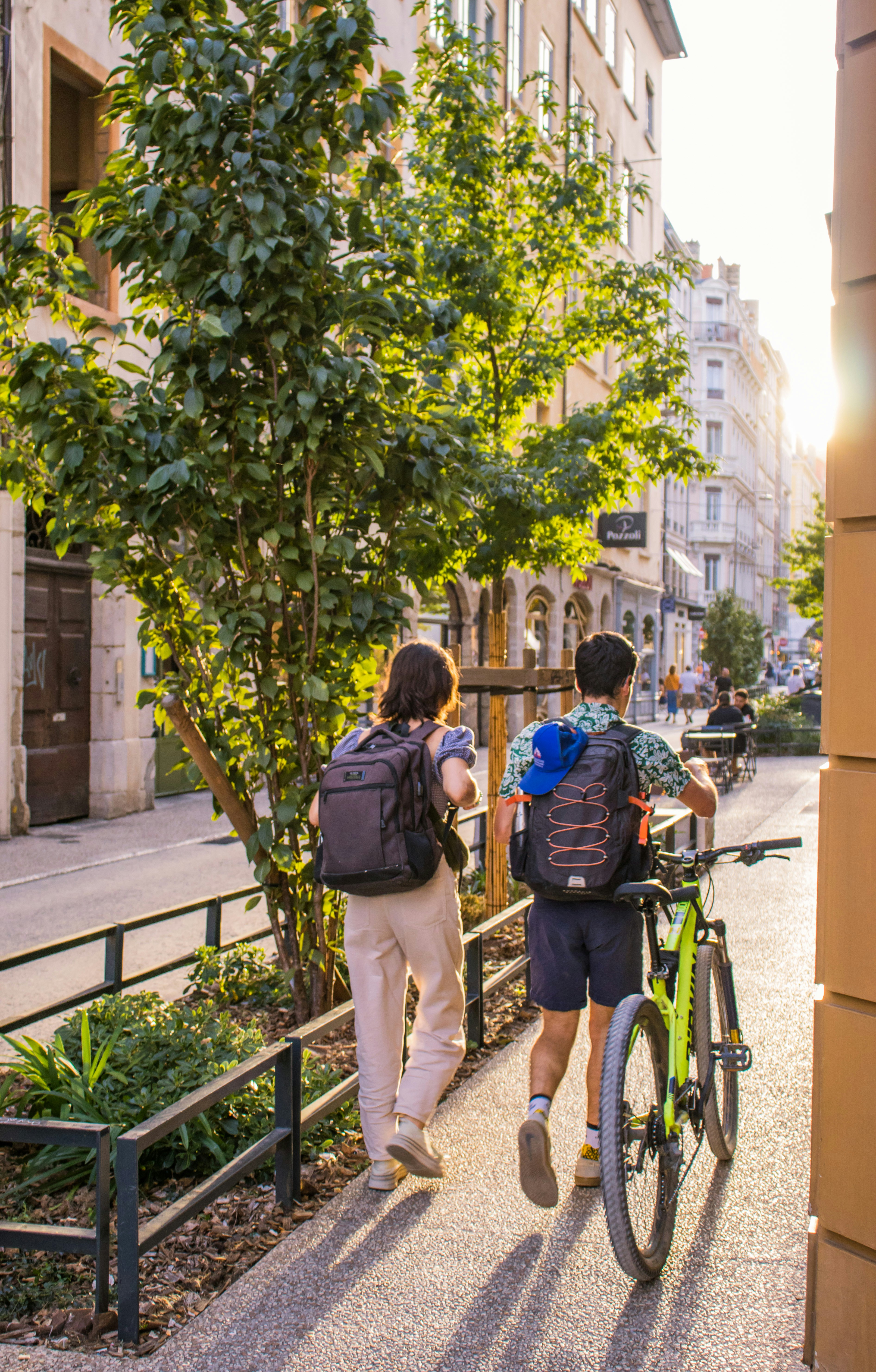 Duas pessoas com mochilas caminhando por uma rua ensolarada da cidade com uma bicicleta