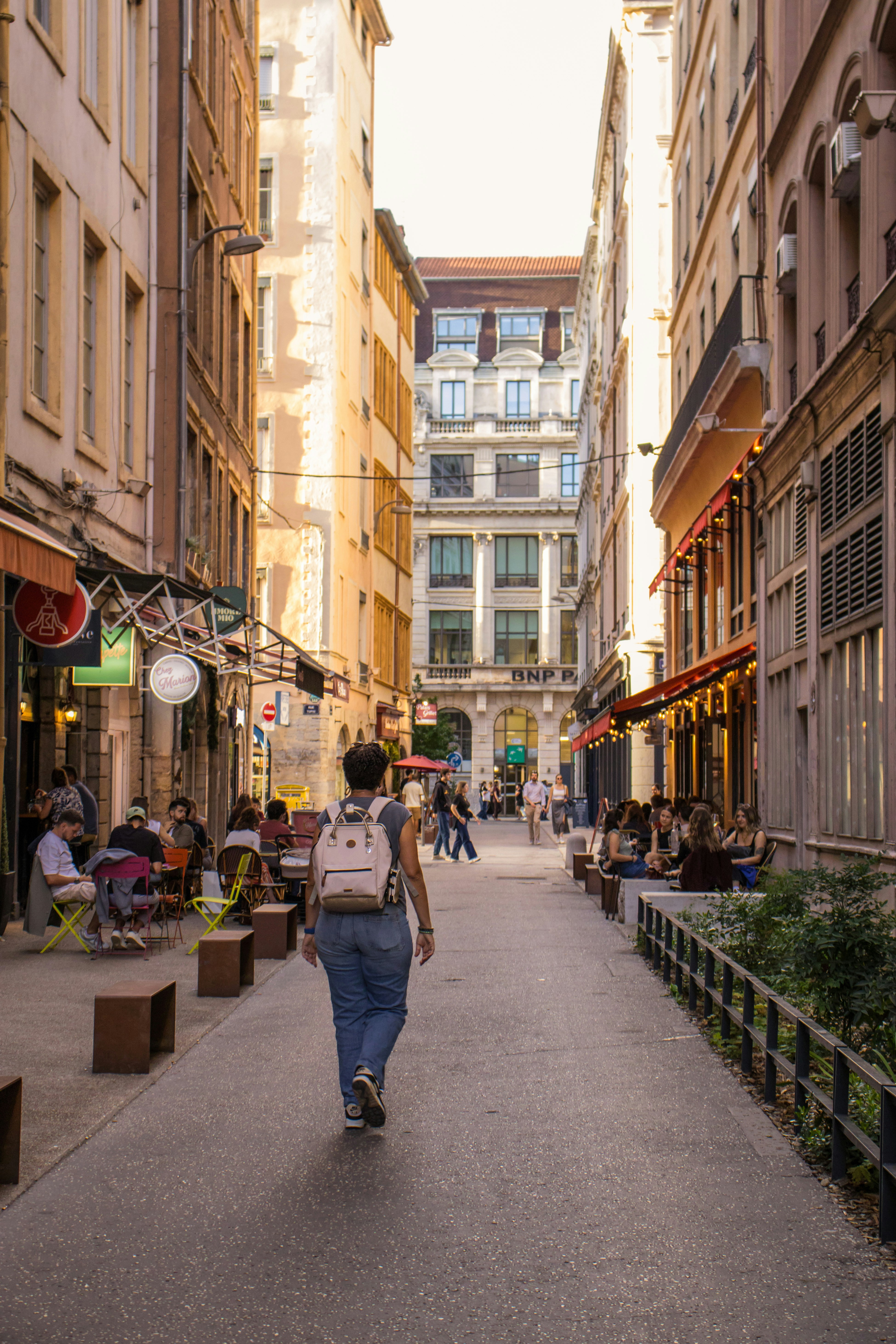 Uma pessoa com uma mochila caminhando por uma rua estreita com assentos ao ar livre em um café