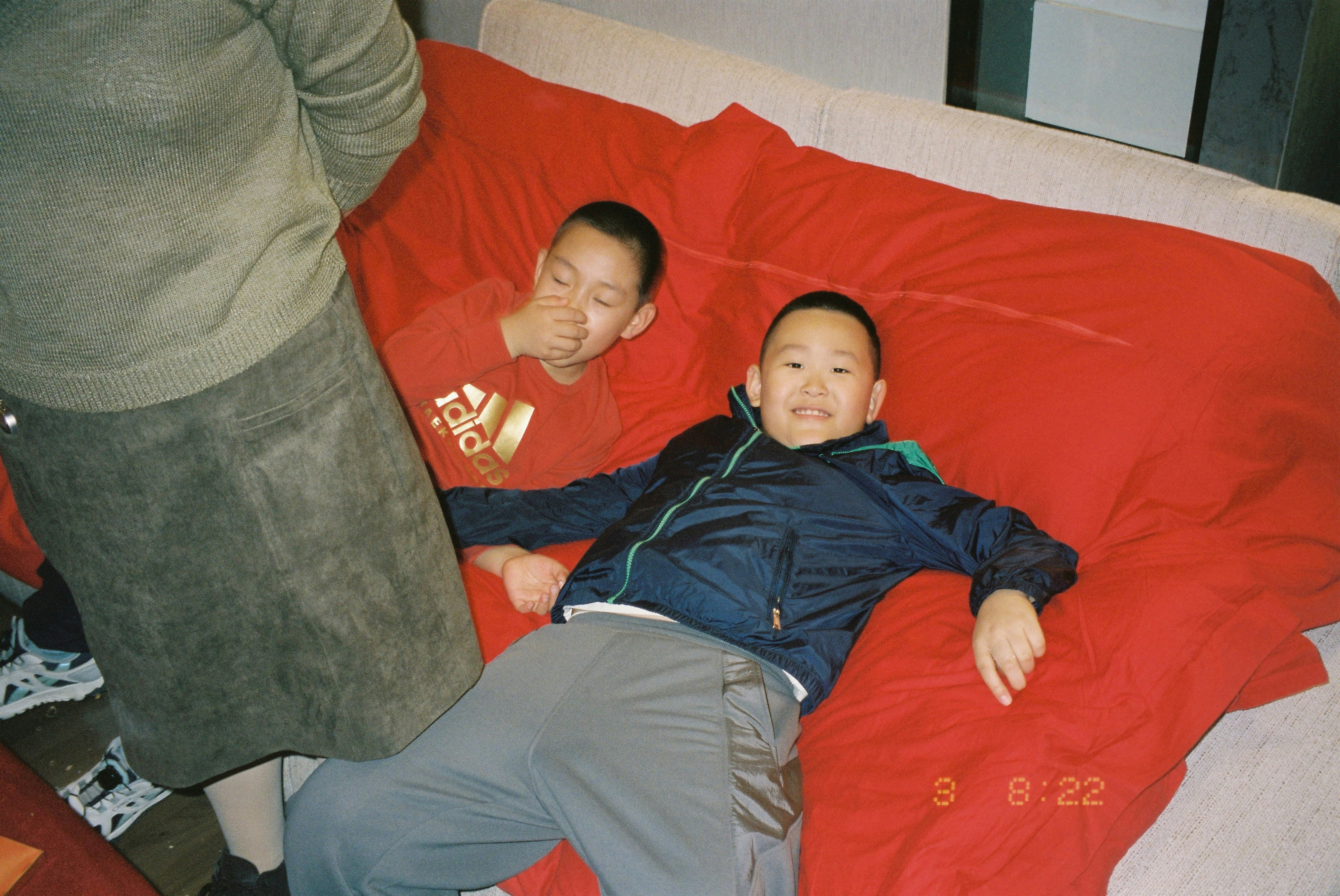 Two children lying on a red sofa beside an adult in a skirt