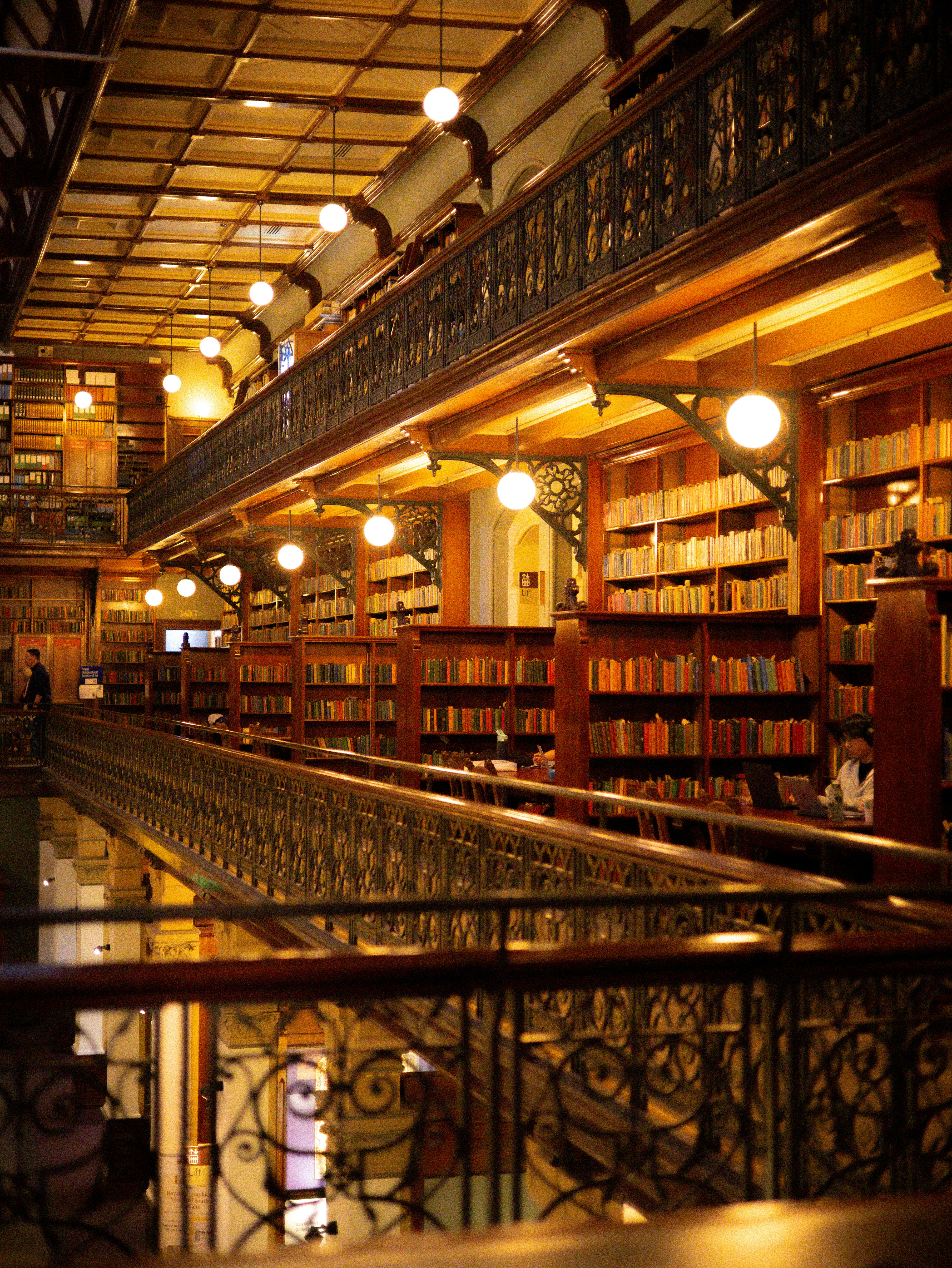 A multi-level library with wooden bookshelves, ornate iron railings, and warm pendant lighting