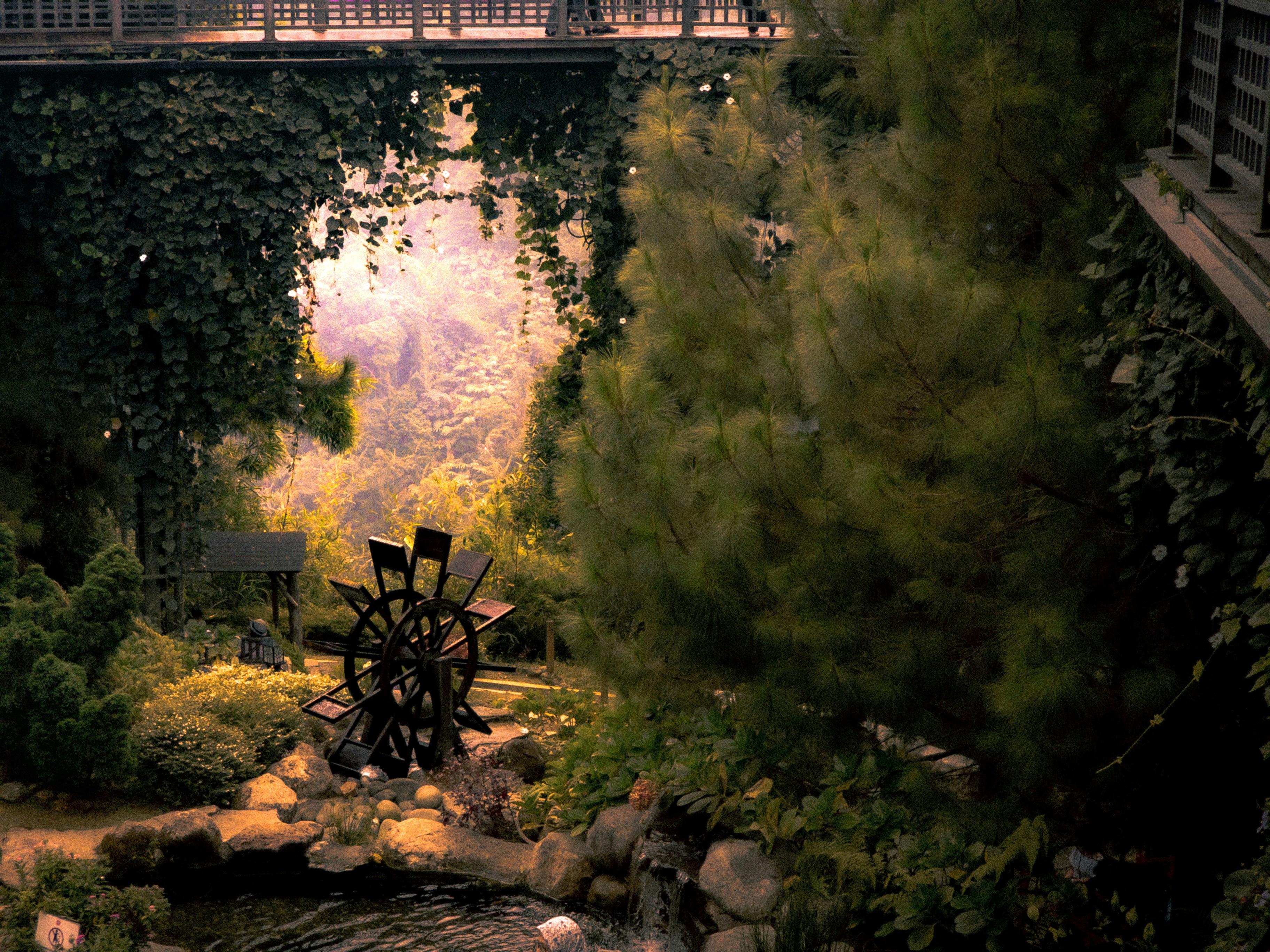 A wooden water wheel in a lush garden with a bridge above