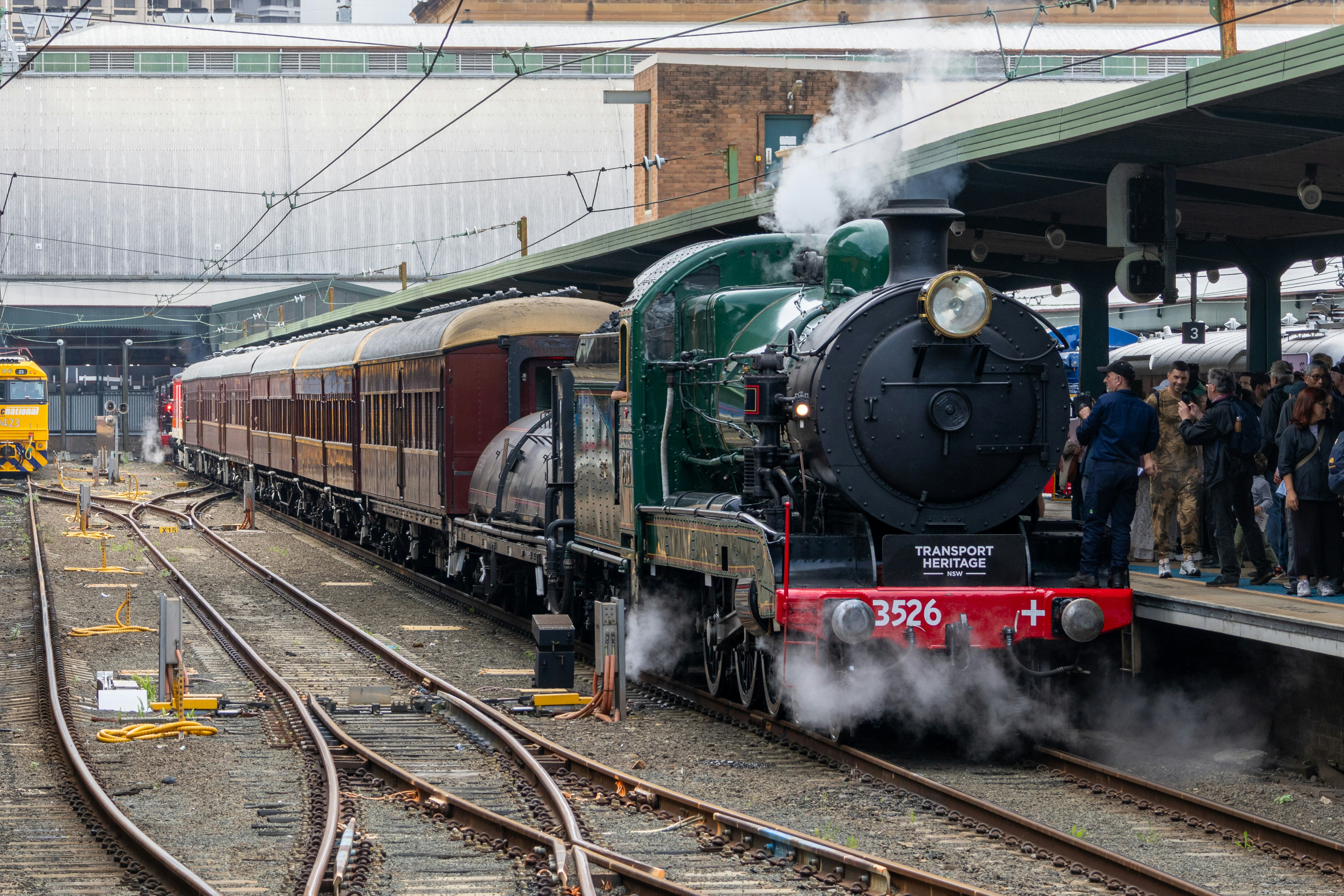 A green steam locomotive pulling vintage passenger carriages at a train station platform