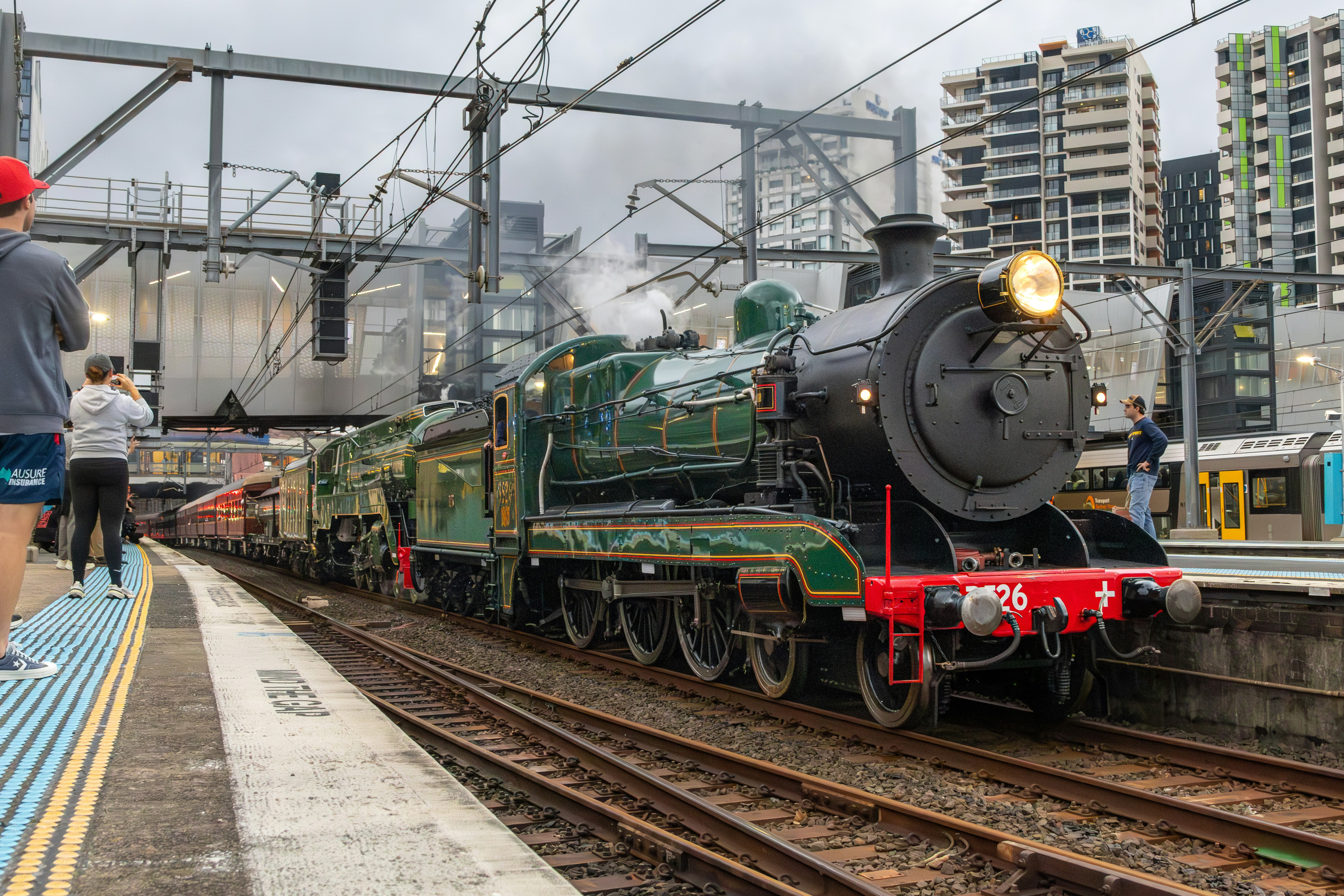 A green steam locomotive pulling passenger carriages into a modern city train station