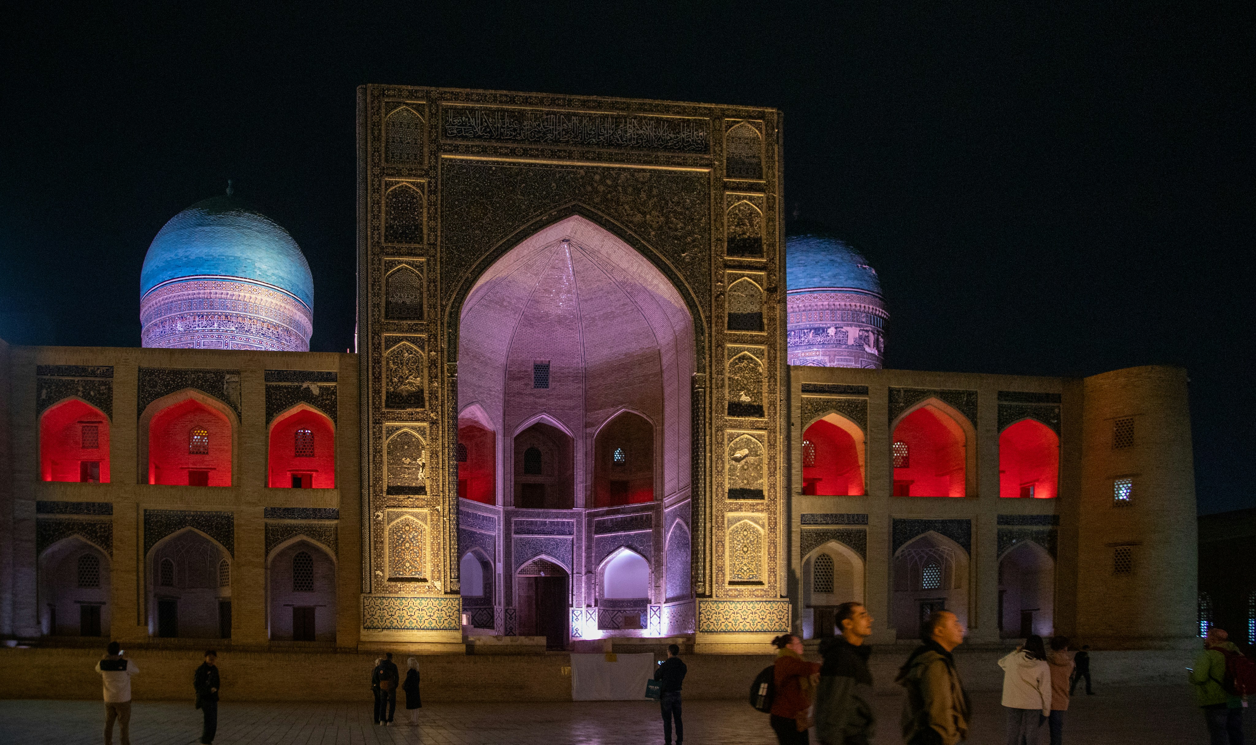 People walking in front of the illuminated Mir-i-Arab Madrasah in Bukhara at night
