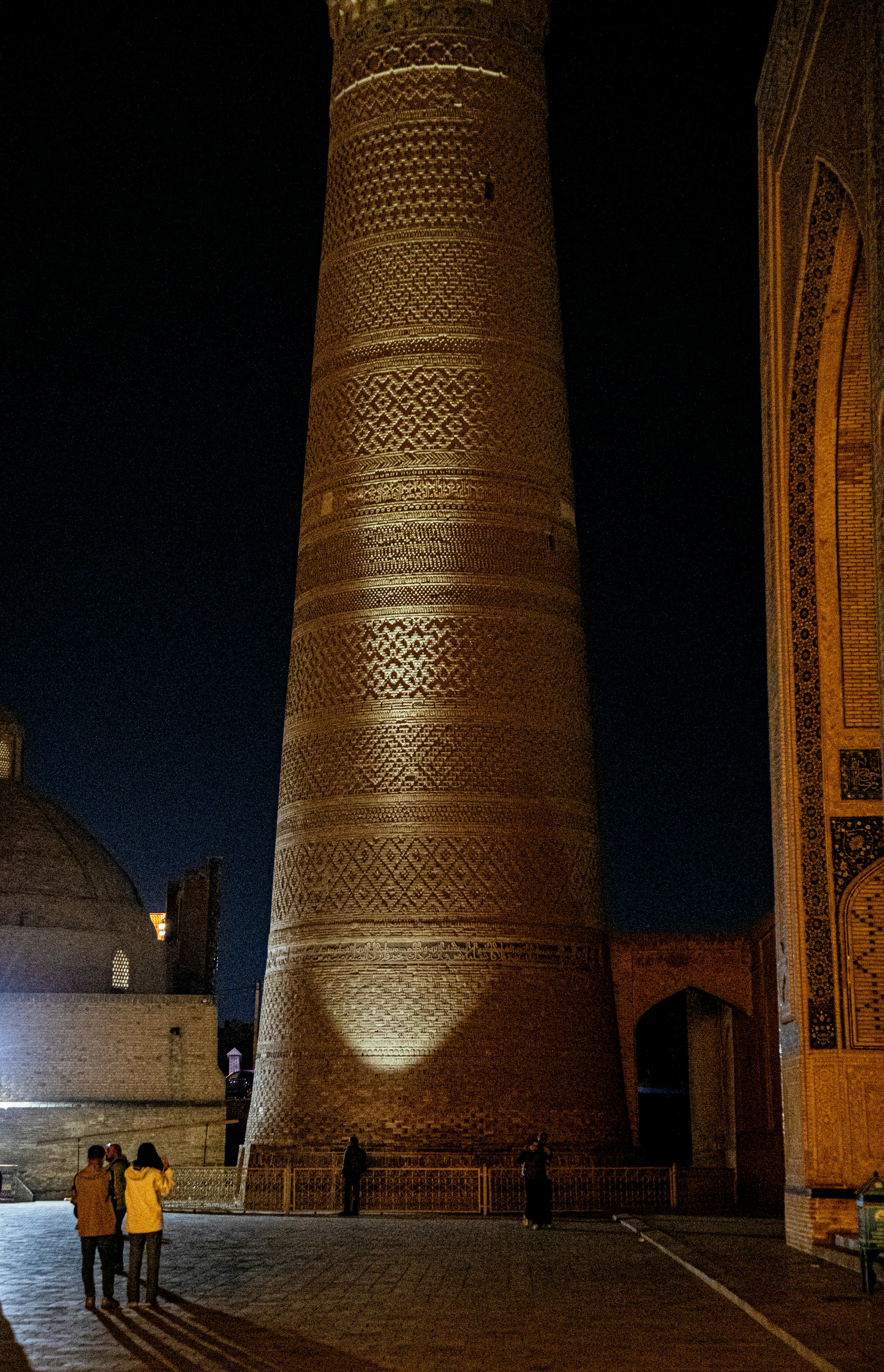 The illuminated Kalyan Minaret at night with people walking in the square in Bukhara