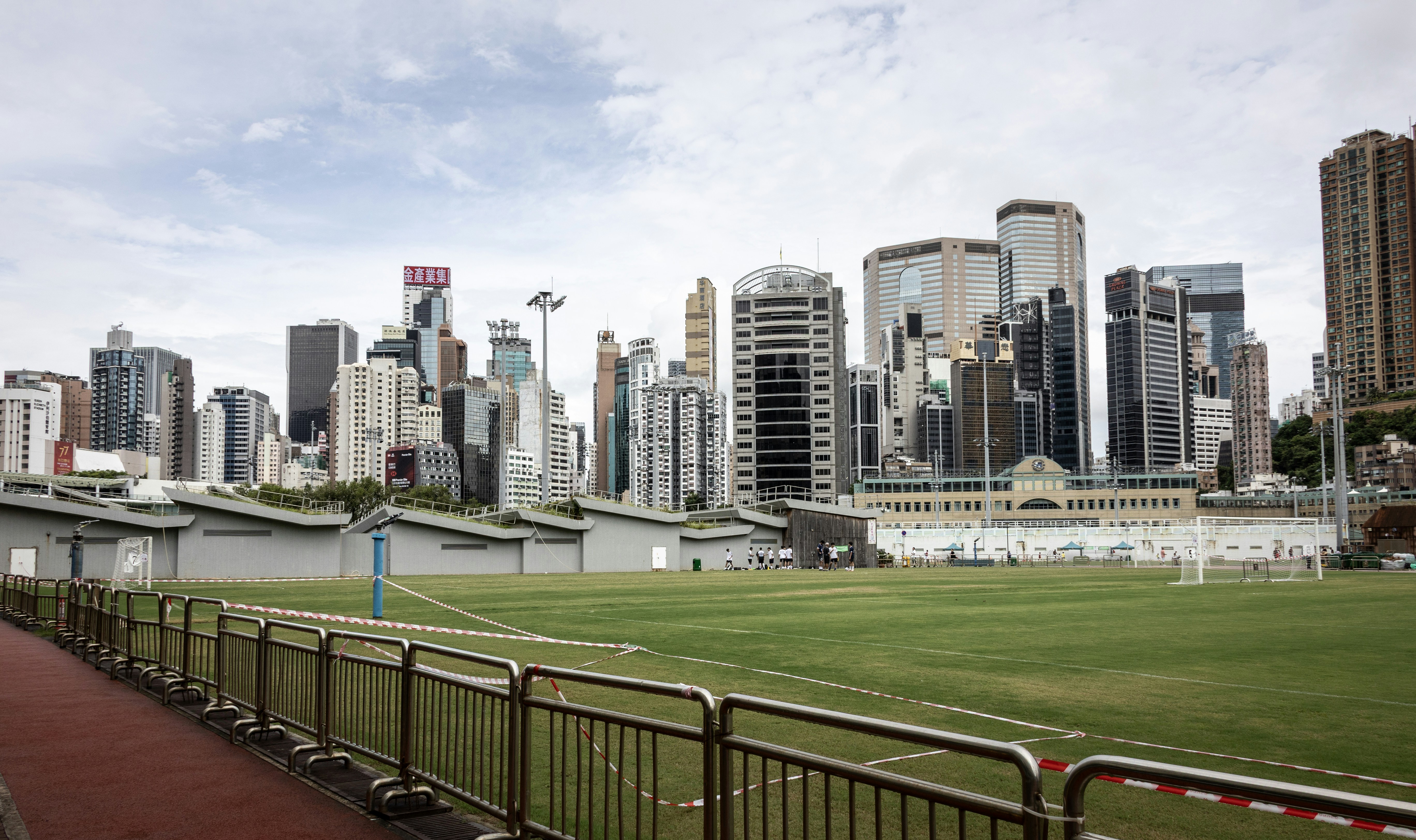 Um campo esportivo verde com barreiras de metal diante de um denso horizonte de Hong Kong
