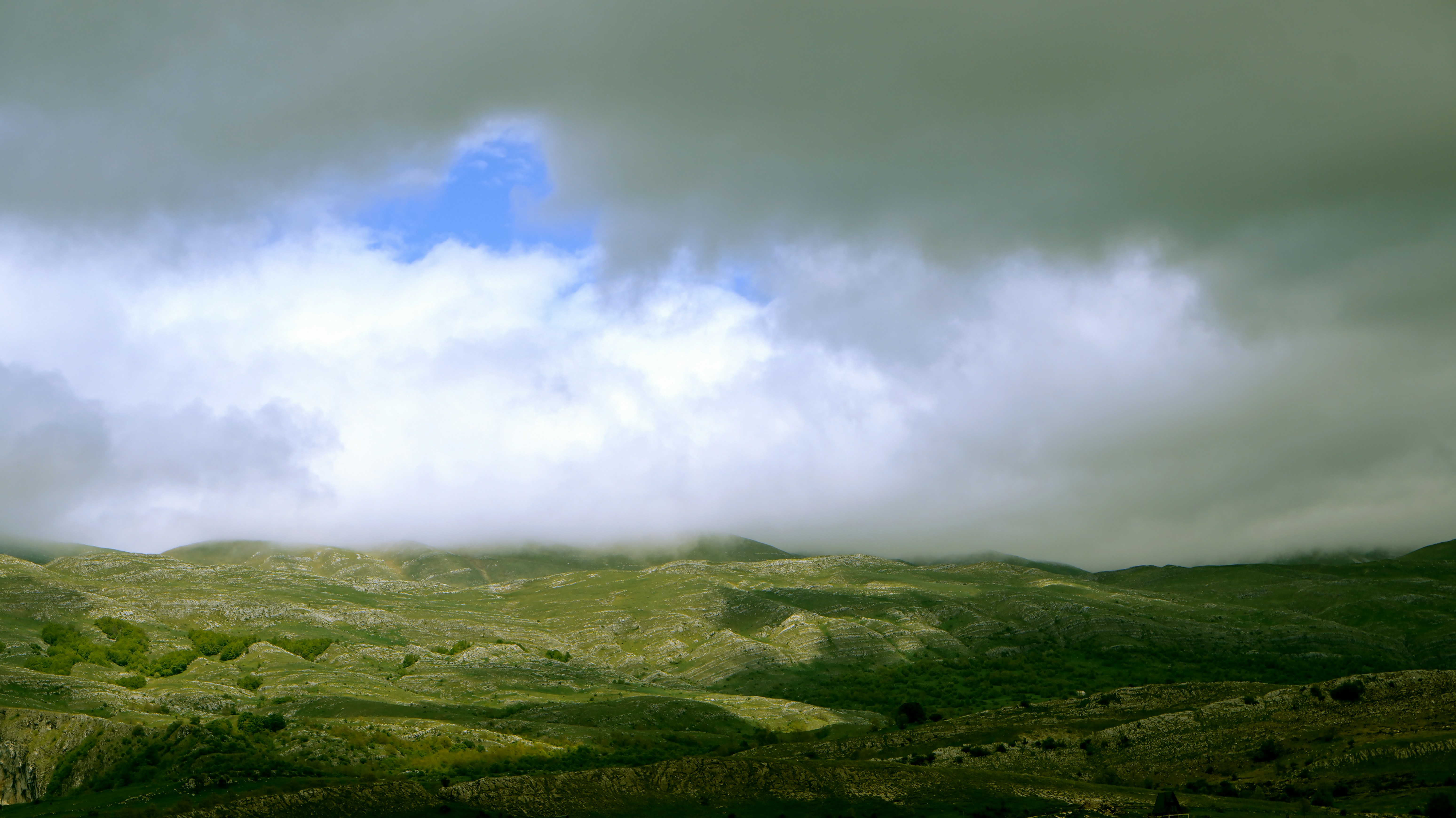 Green rolling hills under a heavy cloud layer with a patch of blue sky