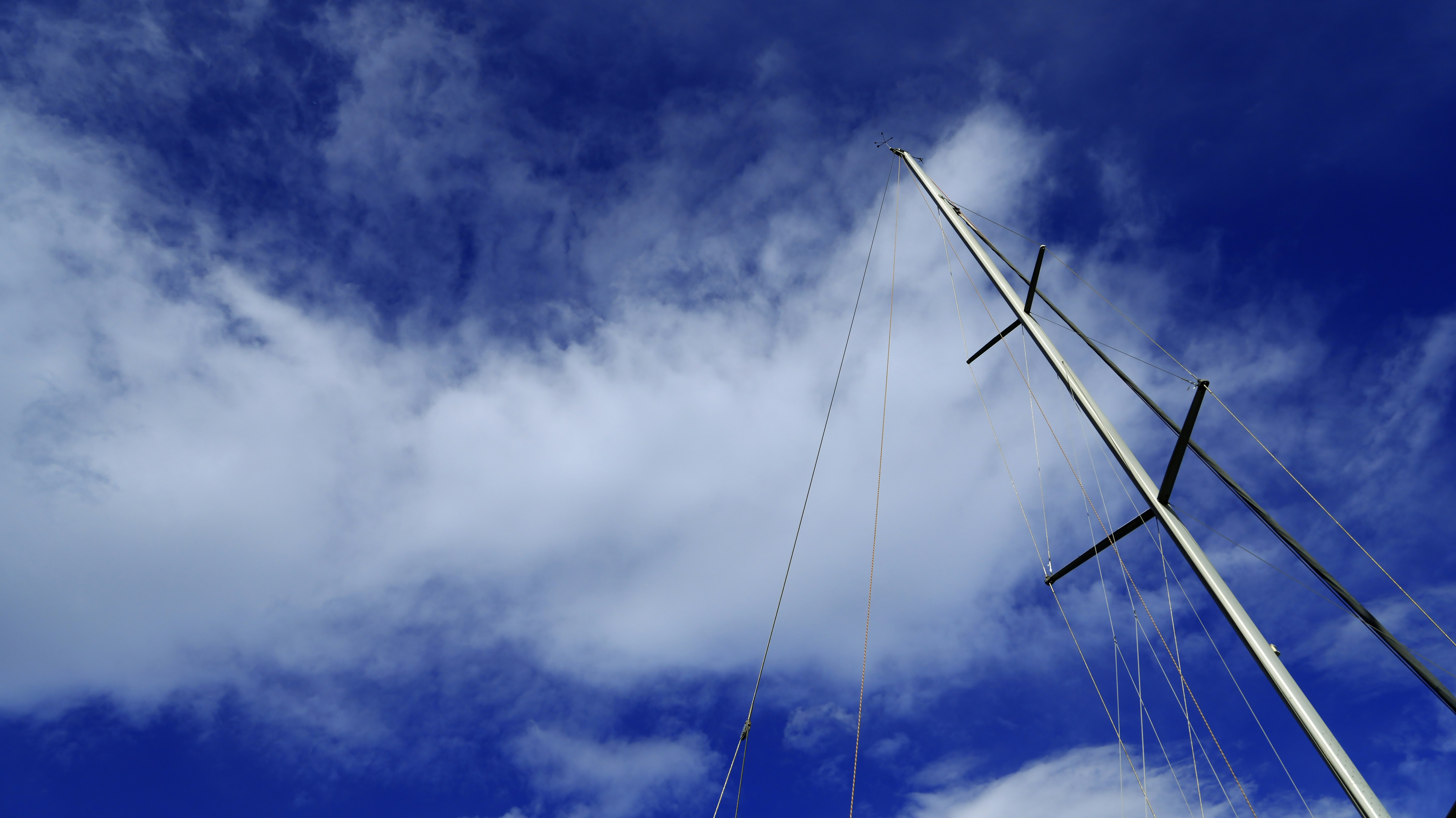 A tall sailboat mast with rigging against a bright blue sky with clouds